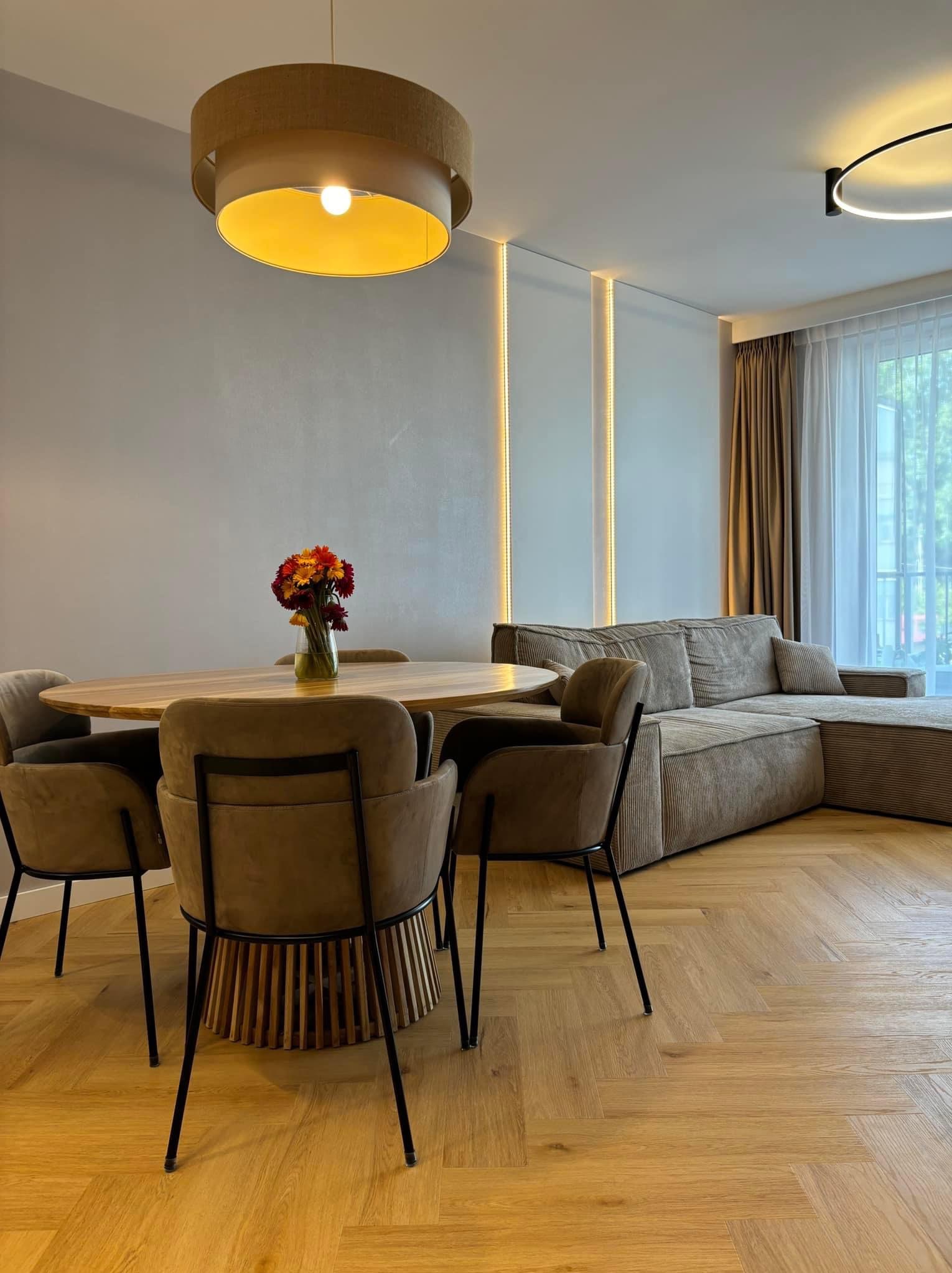 Jasne wnętrze: okrągły stół z bukietem kwiatów, krzesła z czarnymi nogami i drewnianą podstawą, narożna sofa z tkaniny w prążki, parkiet w jodełkę, lampa sufitowa w dwóch odcieniach brązu...