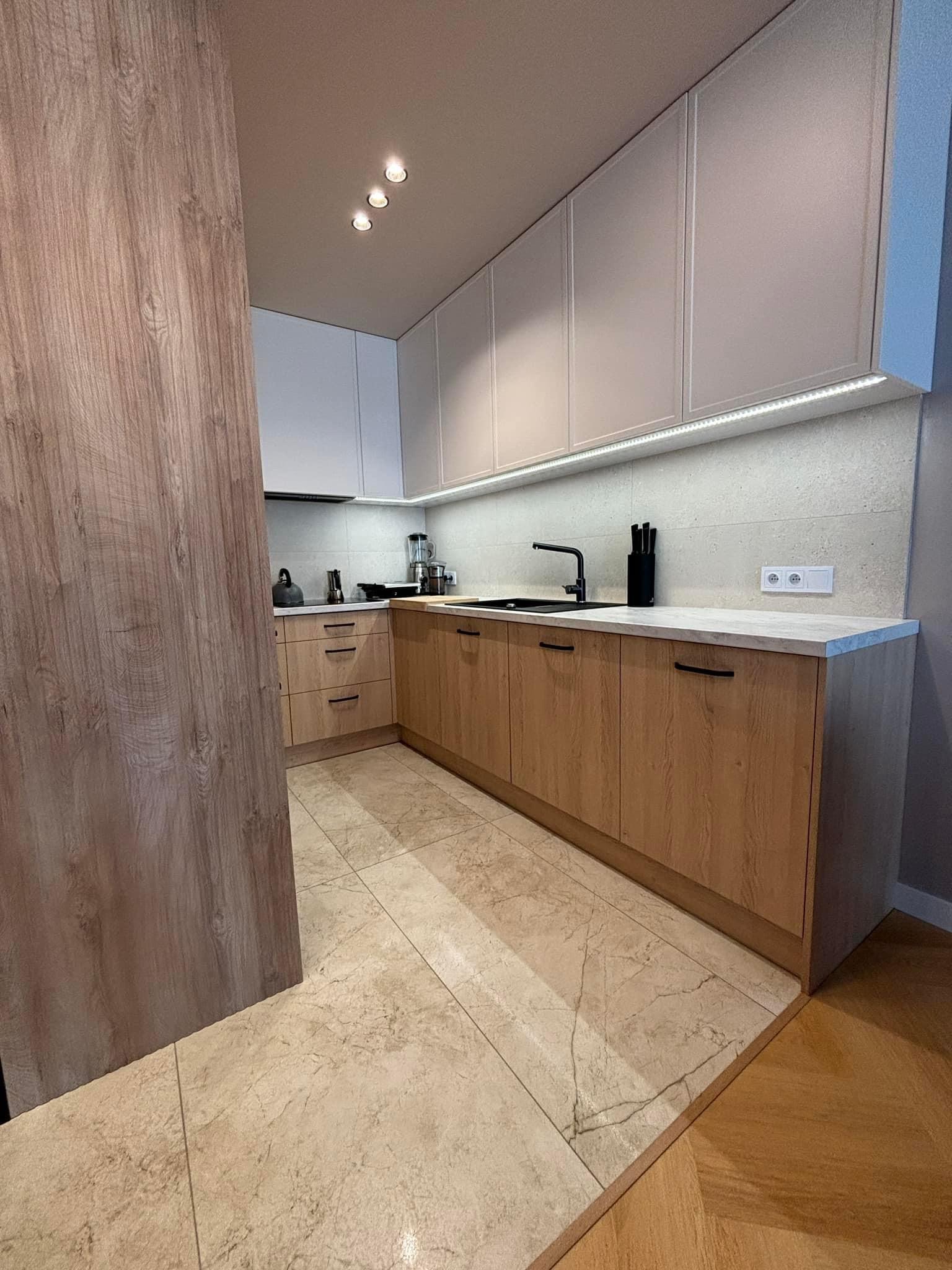 Nowoczesna kuchnia z drewnianymi frontami szafek dolnych i białymi górnymi, oświetleniem LED podszafkowym i beżową podłogą z płytek imitujących marmur, widok zza drewnianej ścianki działowej.