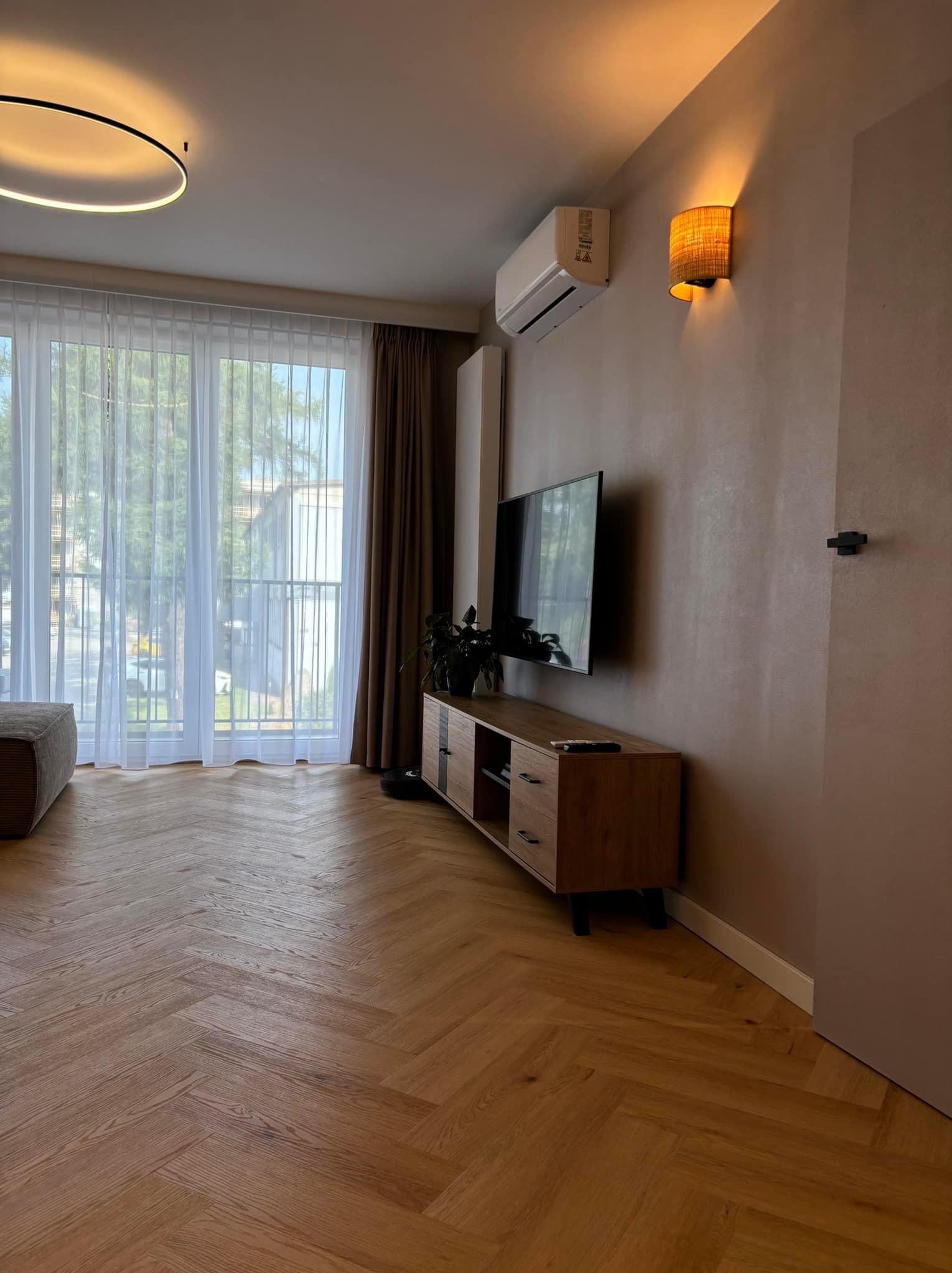 Jasny salon z parkietem w jodełkę, brązowymi ścianami, drewnianą szafką RTV, telewizorem i klimatyzatorem pod sufitem, oświetlony lampą ścienną z abażurem.