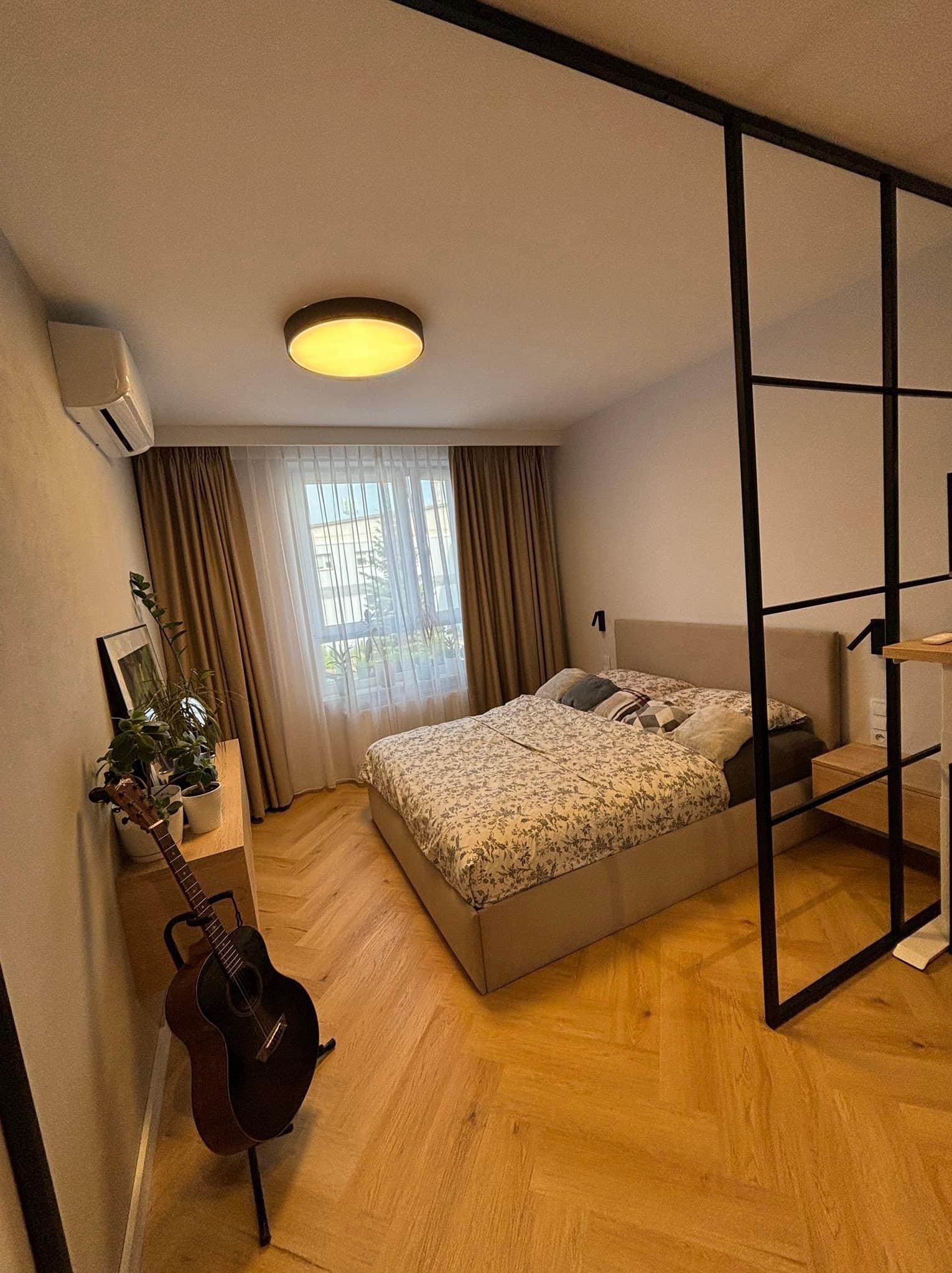 Jasna sypialnia z łóżkiem w kwiaty, drewnianą podłogą ułożoną w jodełkę, czarną ścianką działową z metalowych profili i gitarą opartą o ścianę