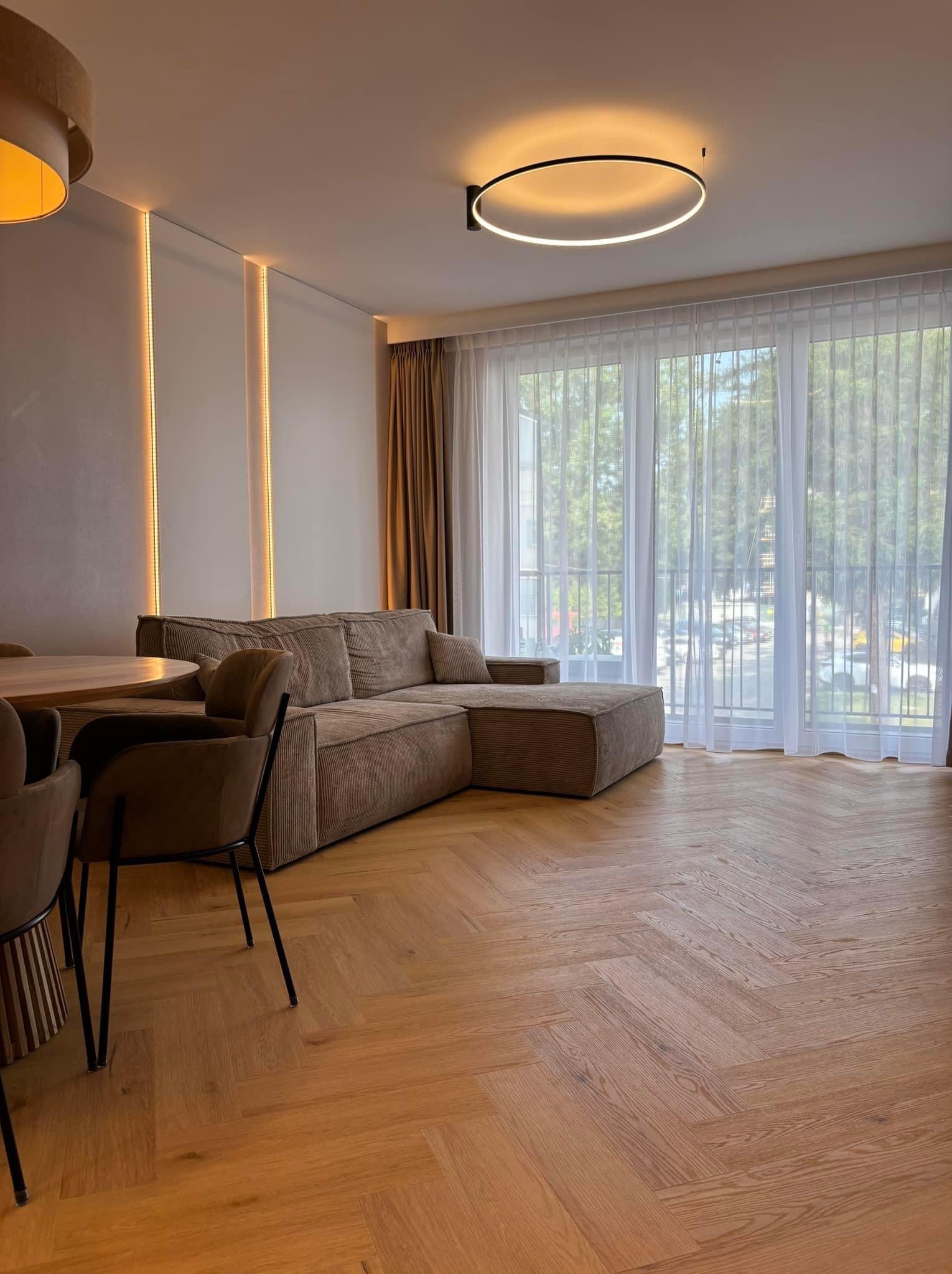 Jasne, przestronne wnętrze salonu z parkietem ułożonym w jodełkę, beżową sofą narożną, okrągłym stołem i krzesłami, oświetlone nowoczesną lampą sufitową i listwami LED na ścianie.