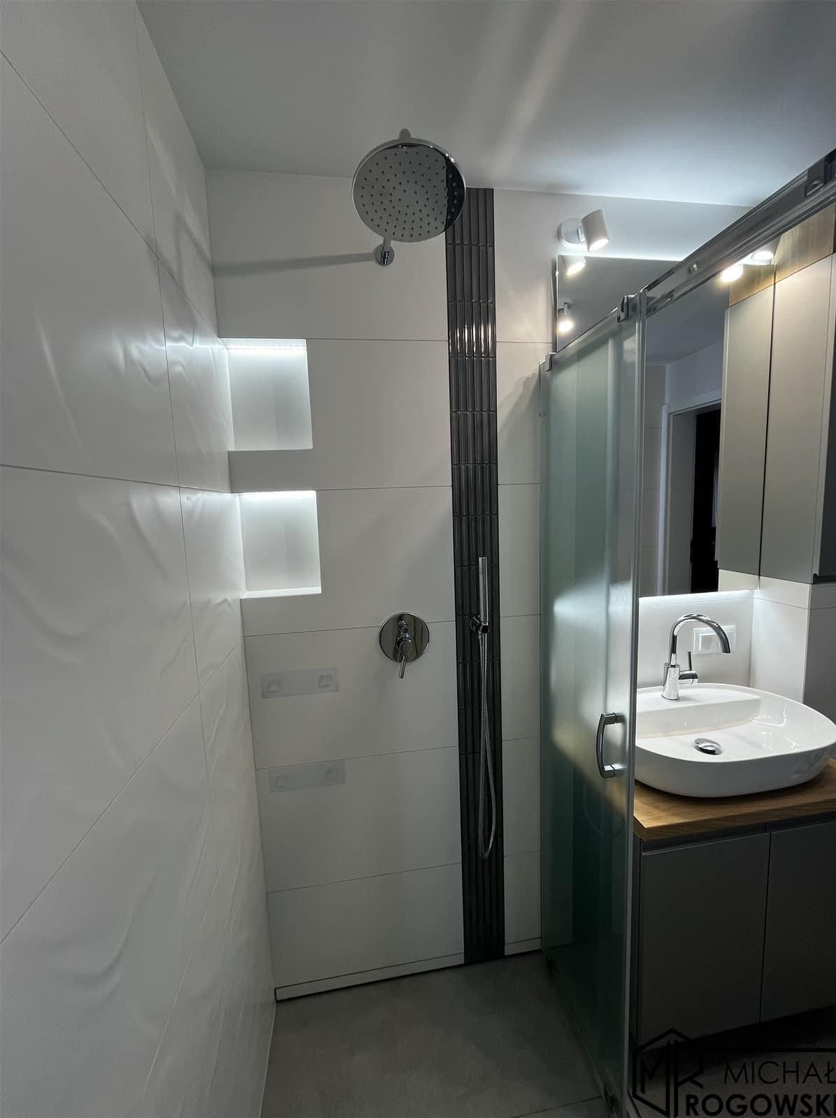 Nowoczesna łazienka z białymi płytkami o falistej strukturze, podświetlanymi wnękami, deszczownicą i szklaną kabiną prysznicową, umywalką nablatową na drewnianym blacie i szarą szafką.