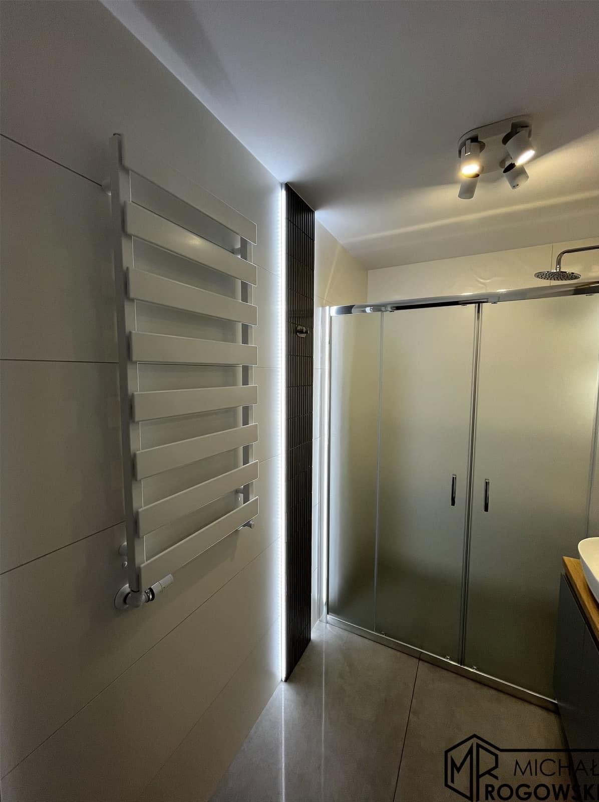 Nowoczesna łazienka z grzejnikiem drabinkowym, kabiną prysznicową z matowego szkła i oświetleniem LED w pionowym panelu dekoracyjnym.