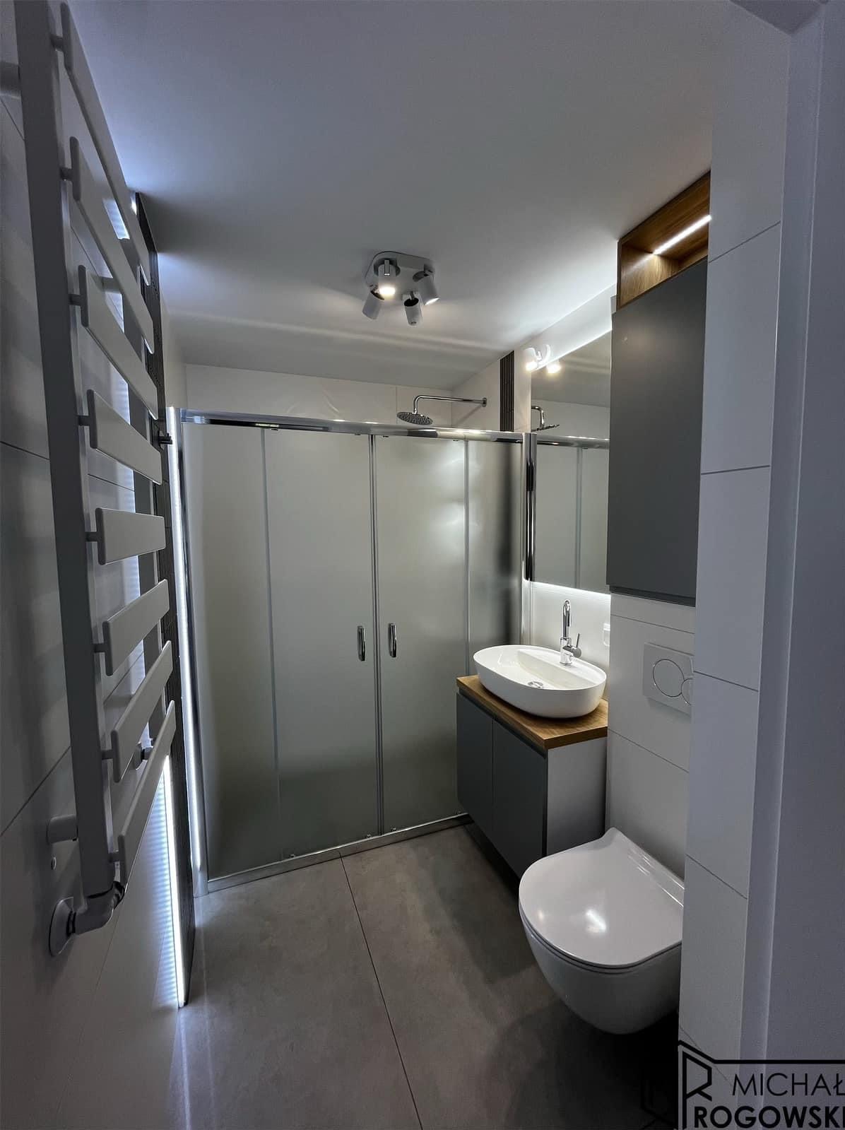 Nowoczesna łazienka z kabiną prysznicową, umywalką nablatową na drewnianym blacie, podświetlanym grzejnikiem drabinkowym i szarą armaturą.