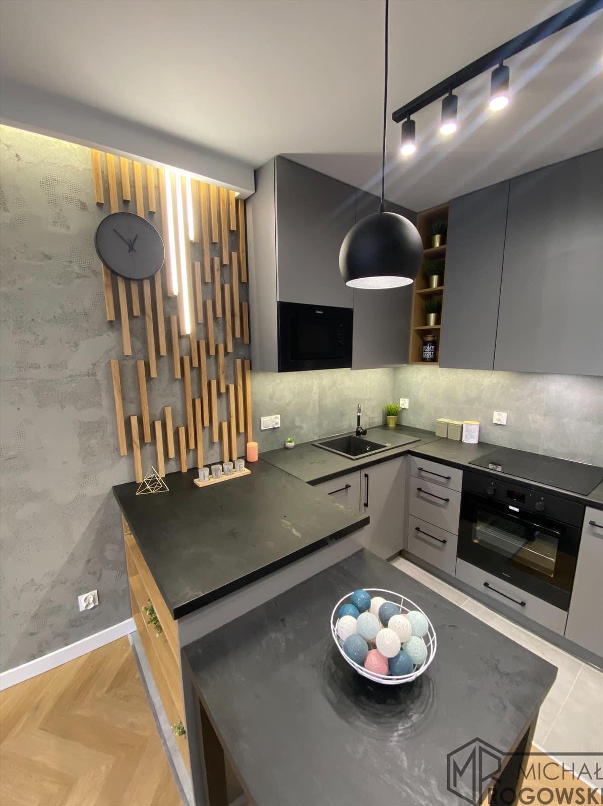 Nowoczesna kuchnia w odcieniach szarości z drewnianymi akcentami dekoracyjnymi na ścianie, oświetleniem punktowym i wyspą z dekoracyjną misą kul.