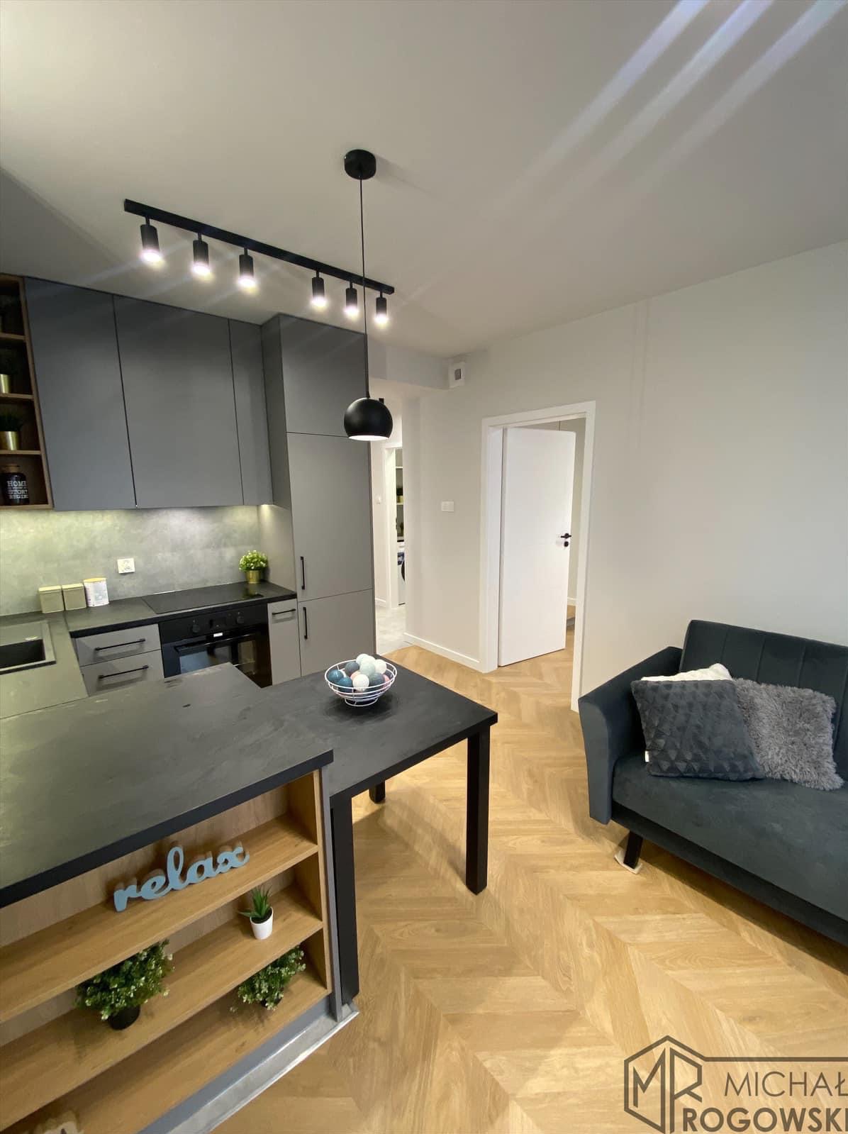 Nowoczesne wnętrze mieszkania z szarą zabudową kuchenną, wyspą z półkami, parkietem w jodełkę i minimalistyczną sofą w tle.