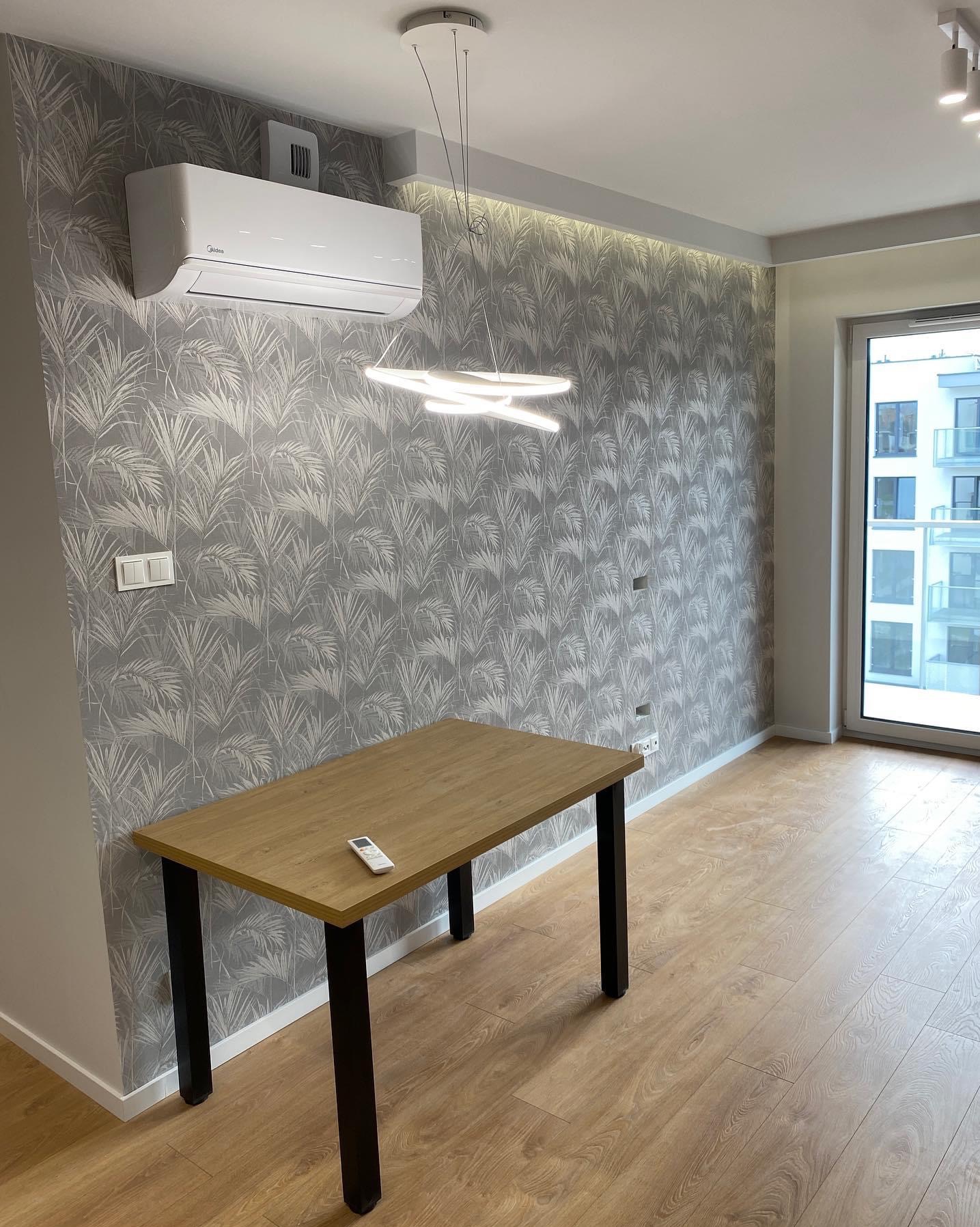 Jasne wnętrze pokoju z drewnianą podłogą, ścianą w tapetę z motywem palm, nowoczesną lampą sufitową i klimatyzatorem Midea, z widokiem na budynek mieszkalny za oknem.