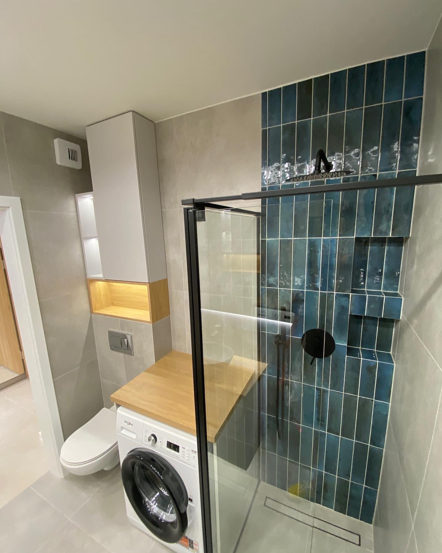 Nowoczesna łazienka z kabiną prysznicową typu walk-in, wykończona błyszczącymi, pionowymi, turkusowymi płytkami, z czarną armaturą i odpływem liniowym. Obok umywalki pralka z blatem z jasnego drewna.
