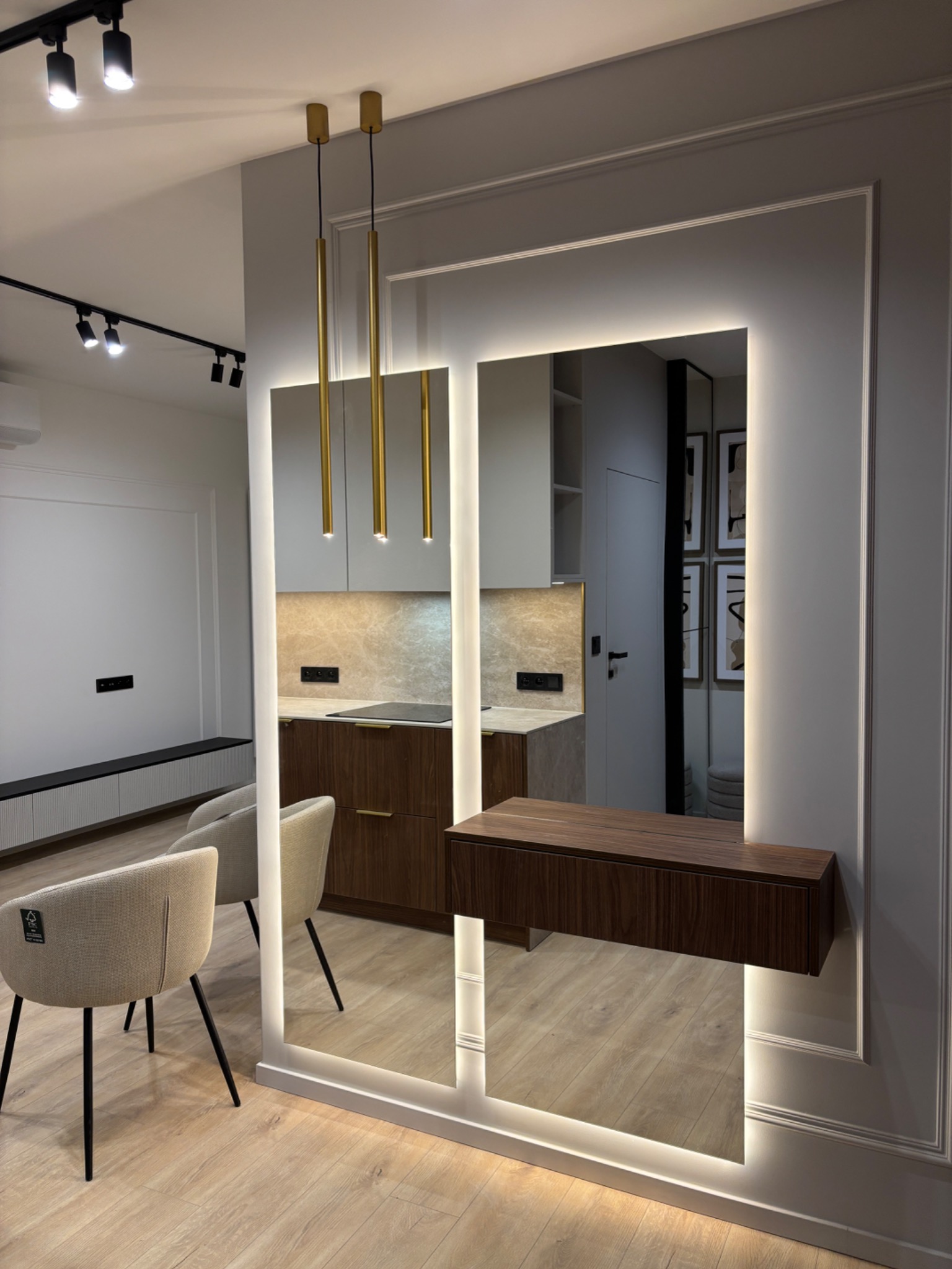 Nowoczesne lustro z podświetleniem LED w ramie, zawieszone na ścianie z dekoracyjnymi listwami, odbijające fragment kuchni z drewnianymi szafkami i krzesła w salonie.