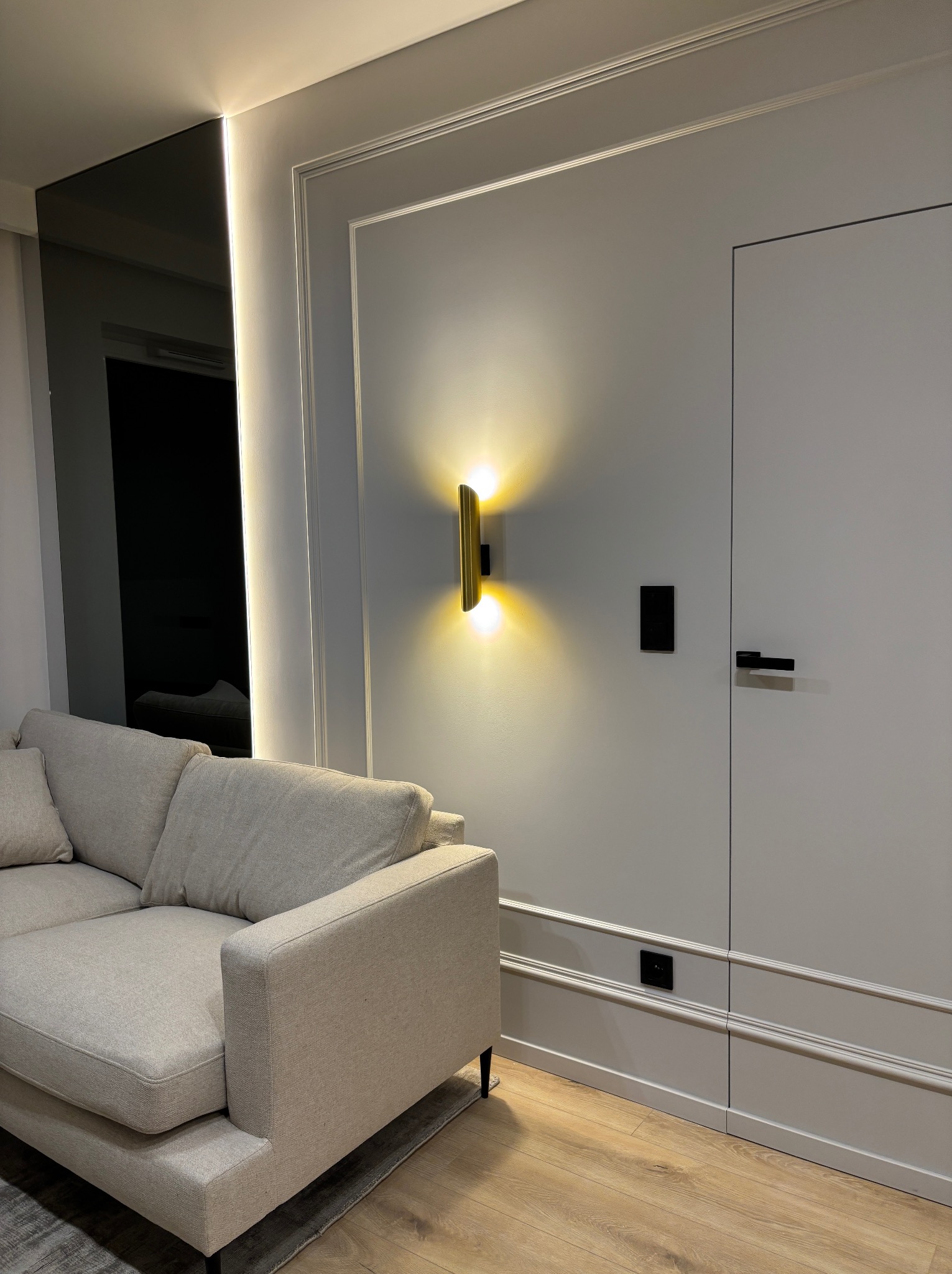 Nowoczesny salon z szarą sofą, ścianą z dekoracyjnymi listwami, złotą lampą ścienną i ukrytymi drzwiami w kolorze ściany.