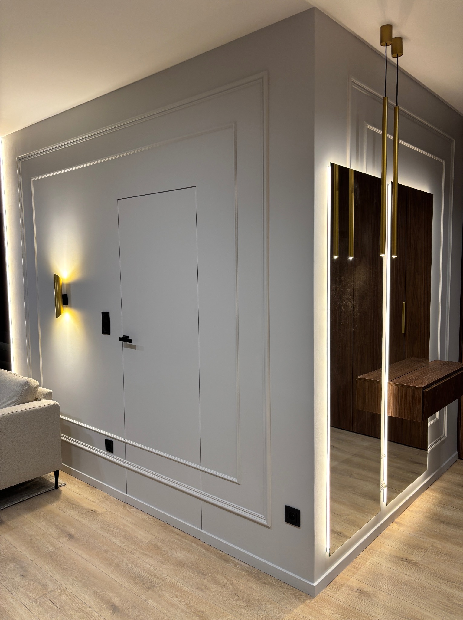 Nowoczesne wnętrze z szarą ścianą ozdobioną sztukaterią, ukrytymi drzwiami, złotym kinkietem i lustrem z podświetleniem LED, odbijającym drewnianą szafę z toaletką.