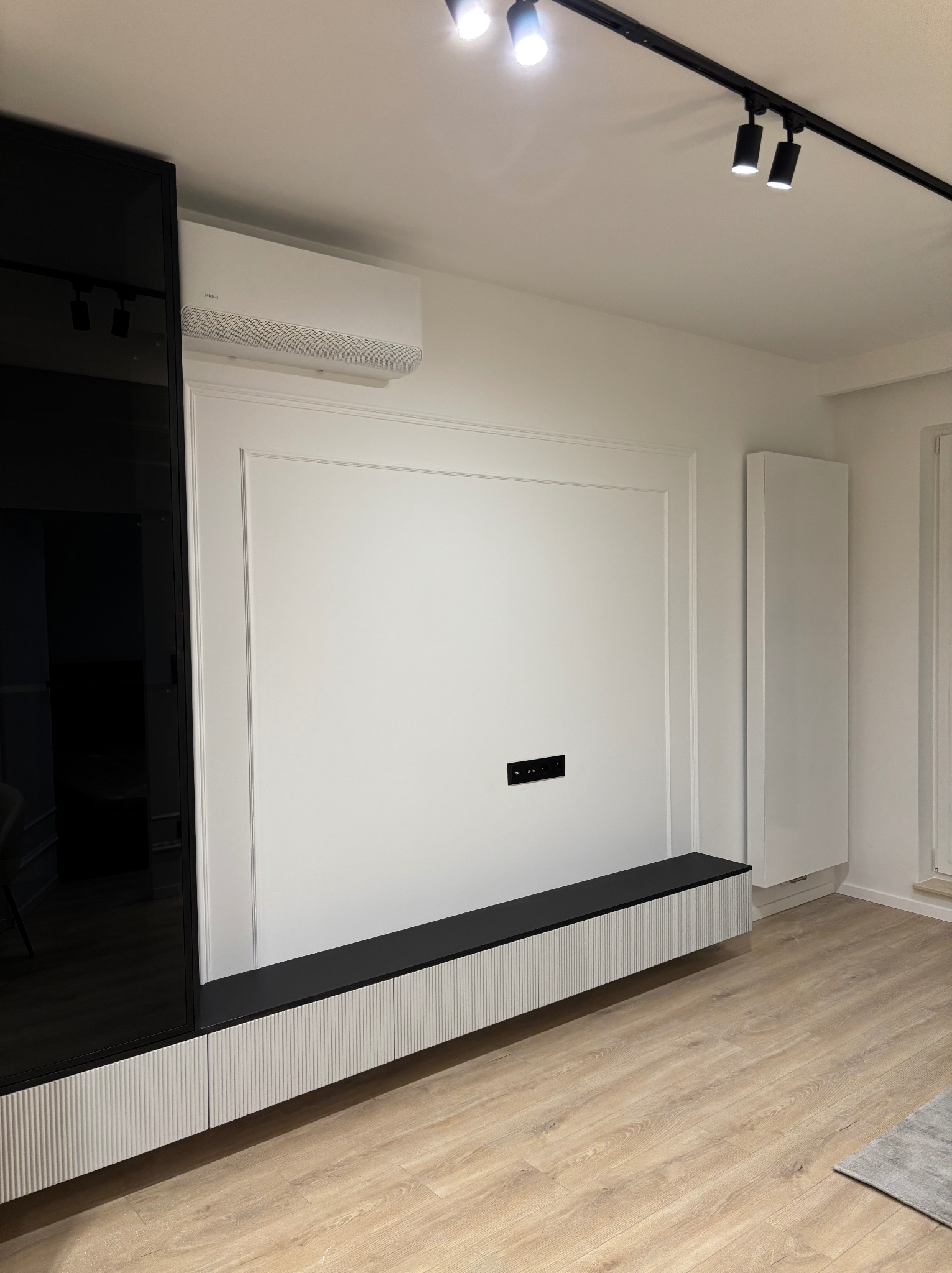 Nowoczesna aranżacja ściany telewizyjnej z czarną szafką RTV, białą ścianą z ozdobnymi listwami, klimatyzatorem i oświetleniem szynowym, na podłodze panele w odcieniach drewna.