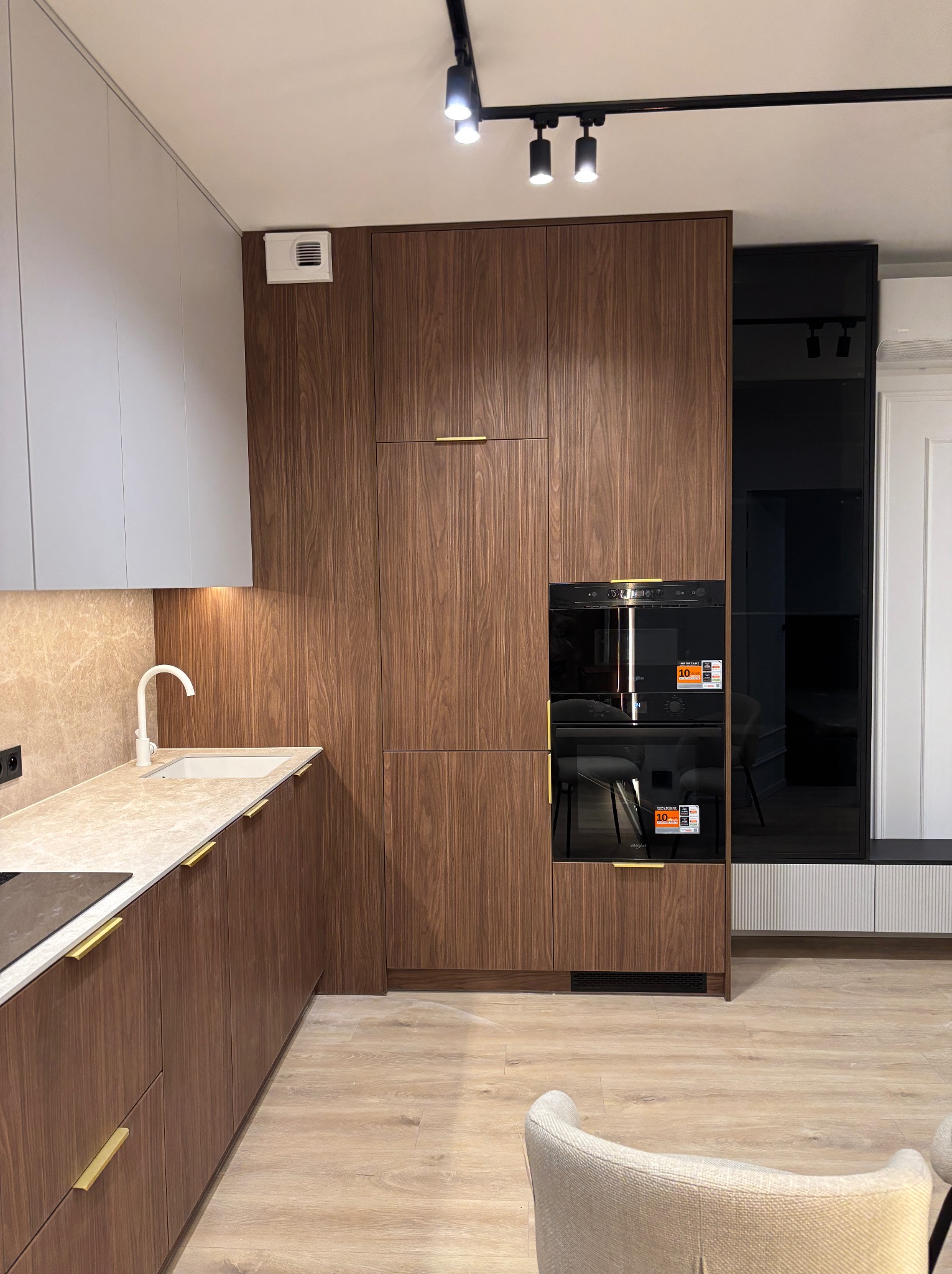 Nowoczesna kuchnia z drewnianymi frontami szafek, białym blatem i złotymi uchwytami, oświetlona reflektorami sufitowymi.