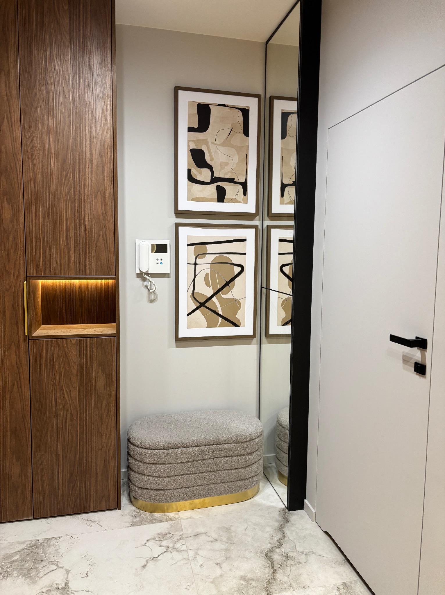 Nowoczesne wykończenie przedpokoju w Krakowie: zabudowa szafy z oświetloną niszą, tapicerowany puf z elementami złota, abstrakcyjne obrazy na ścianie i duże lustro.