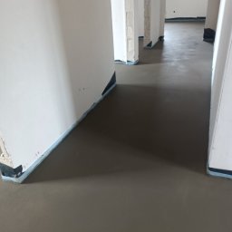GŁOWSKI NIEDZIELSKI SPÓŁKA CYWILNA - Świeżo wylana, gładka posadzka w surowym stanie, z widocznymi dylatacjami przy ścianach, przygotowana do dalszych prac wykończeniowych.