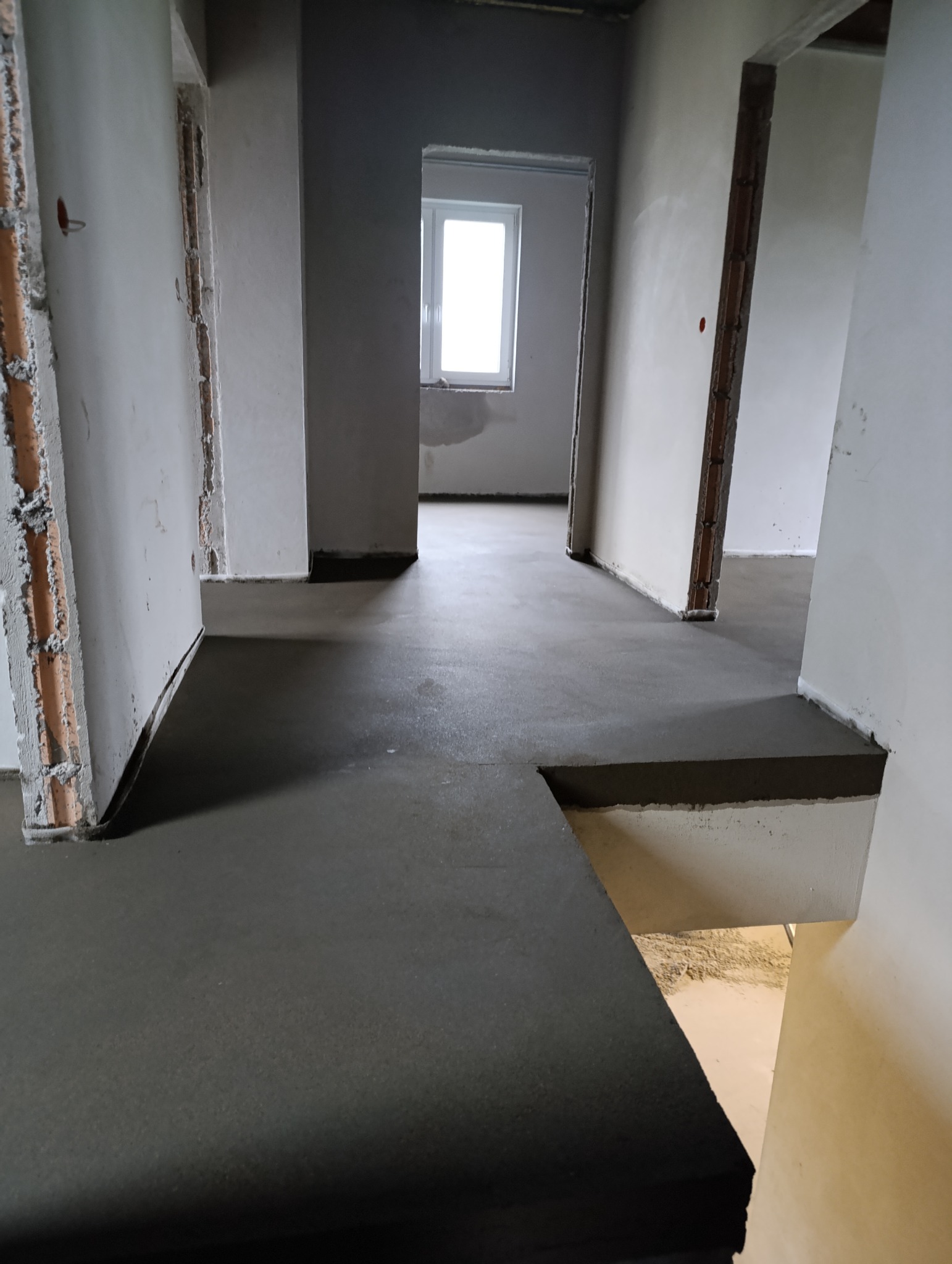 Świeżo wylana, gładka, szara wylewka na podłodze w surowym stanie, widoczne niedokończone ściany i przejścia do innych pomieszczeń.