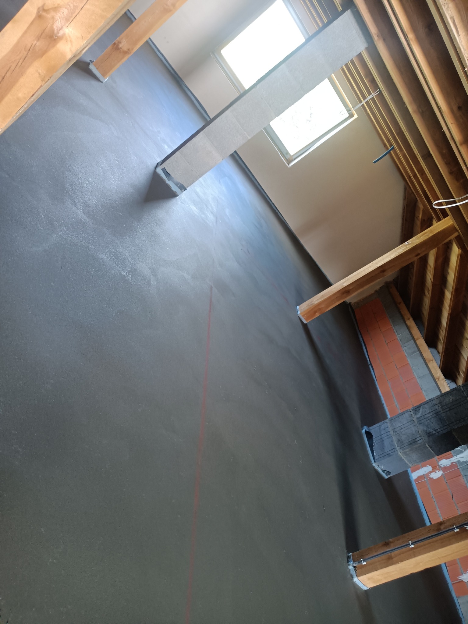 Świeżo wylana, gładka, szara posadzka w surowym wnętrzu poddasza z widocznymi elementami konstrukcji drewnianej i oknem dachowym.