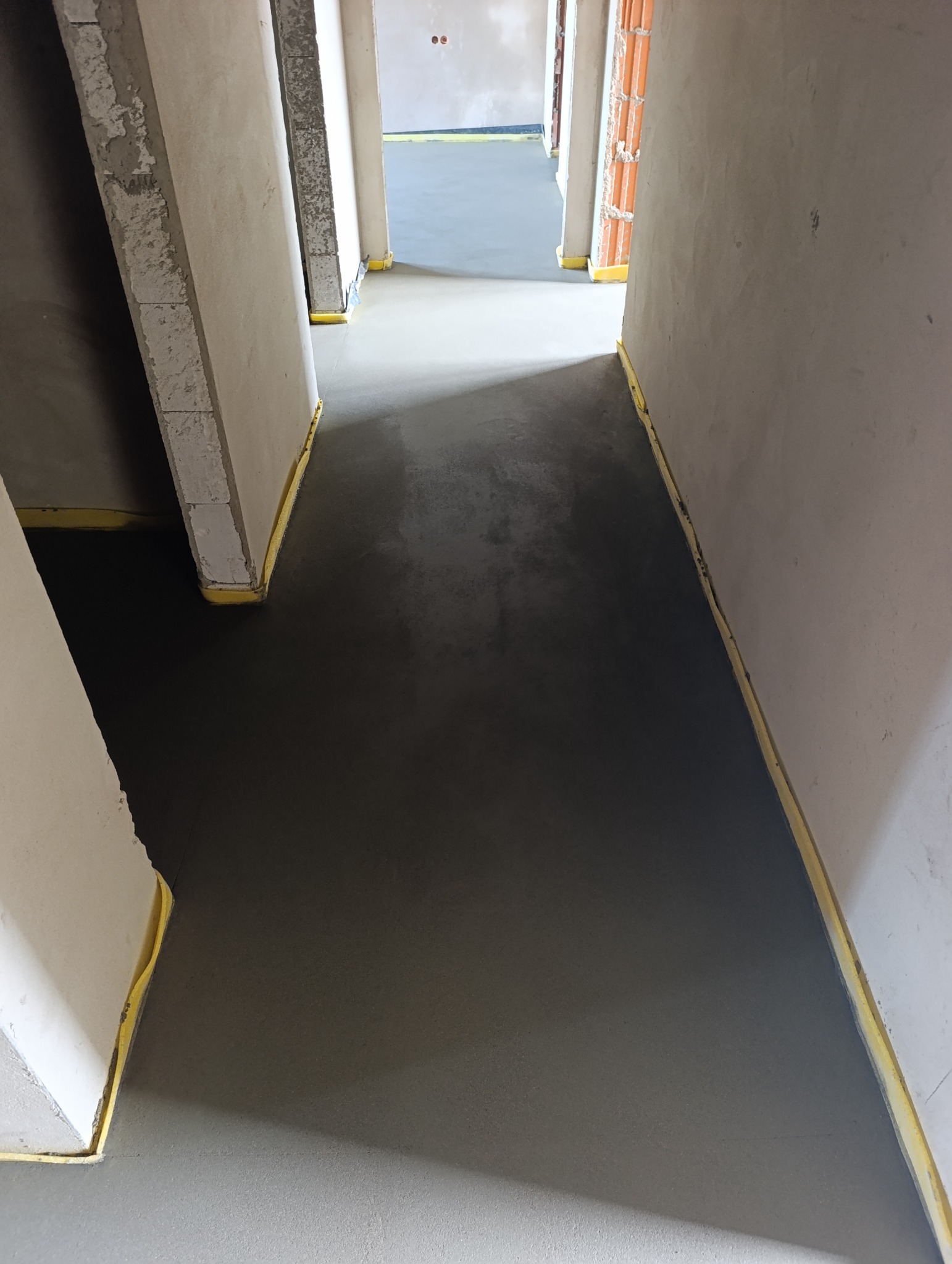 Świeżo wylana, gładka posadzka jastrychowa w korytarzu, zabezpieczona żółtą taśmą przy ścianach. Widok w głąb pomieszczeń, surowy stan wykończenia.