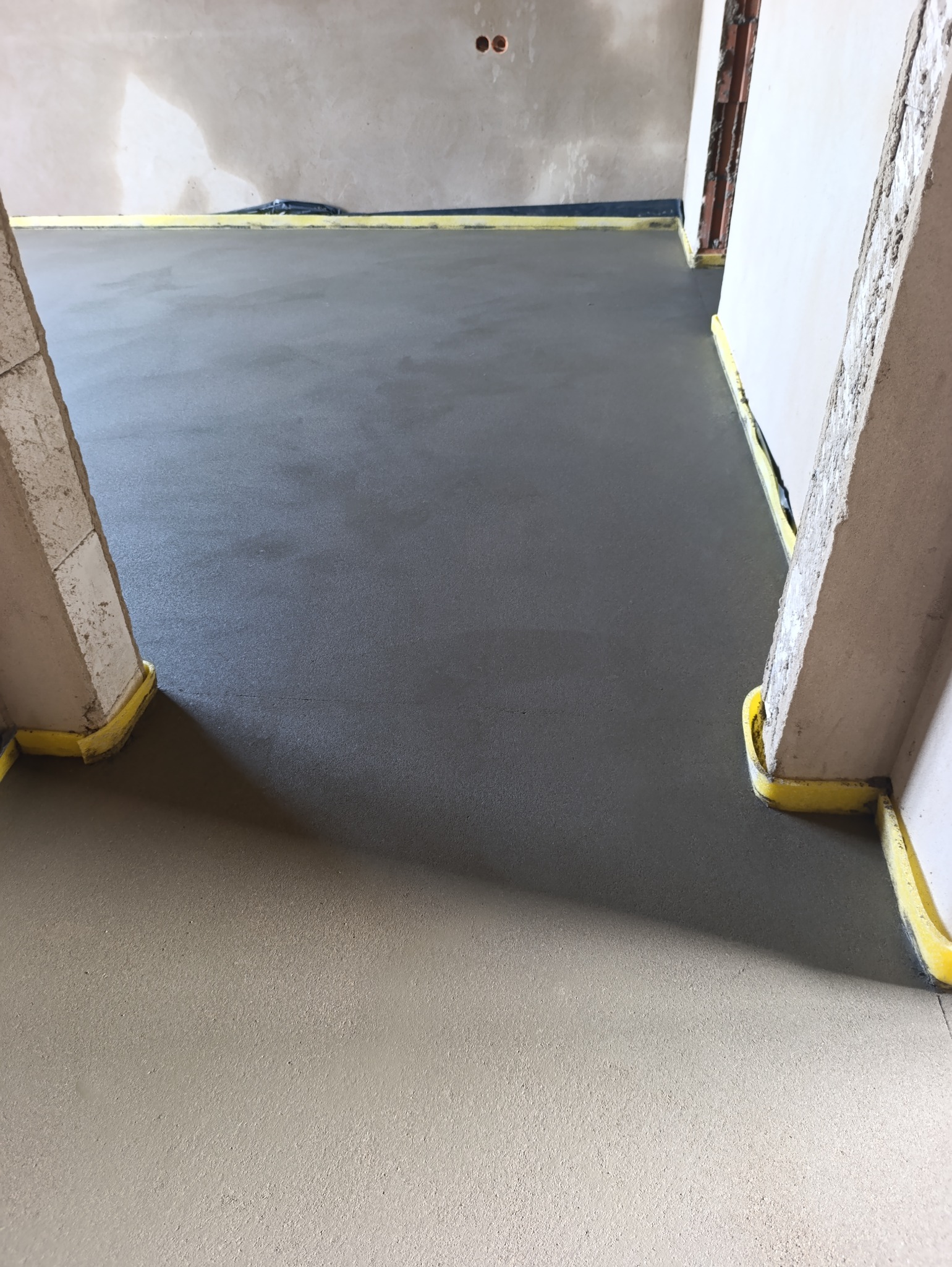 Świeżo wylana, gładka posadzka jastrychowa w pomieszczeniu w trakcie remontu, zabezpieczona żółtą taśmą przy ścianach i framugach drzwi.