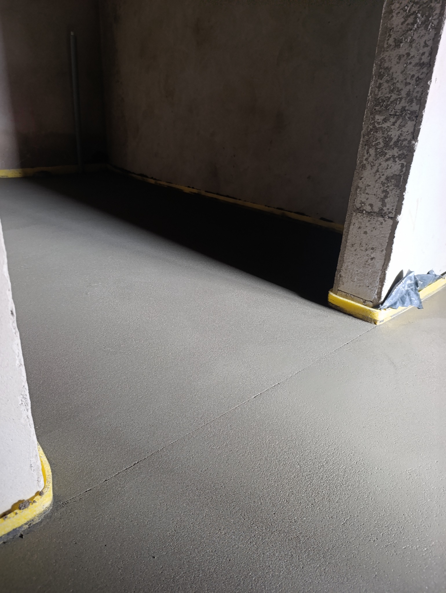 Świeżo wylana, gładka posadzka jastrychowa z żółtą taśmą dylatacyjną przy ścianach w surowym wnętrzu. Widoczna szczelina dylatacyjna.