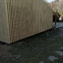 Nowoczesna elewacja z pionowych, drewnianych lameli, tworząca ażurową osłonę na schodach zewnętrznych domu. Surowy stan drewna, widoczne niedoskonałości trawnika.