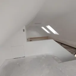 Poddasze po wykonaniu gładzi i malowaniu na biało, widoczne okna dachowe, otwór rewizyjny w ścianie oraz puszki elektryczne bez osprzętu.