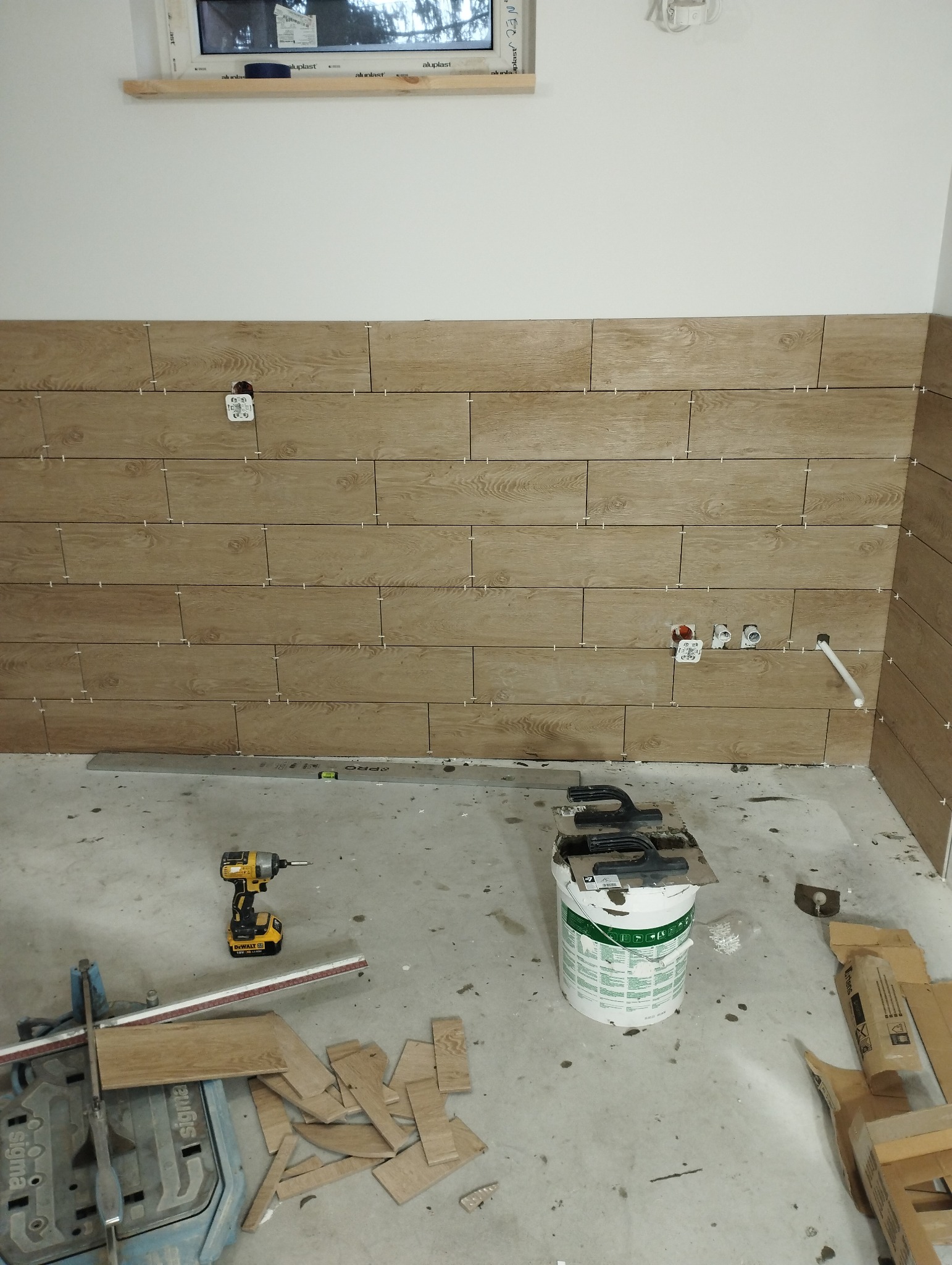 Ściana w trakcie układania płytek imitujących drewno, widoczne puszki elektryczne i rury, narzędzia glazurnicze, poziomica i wiadro z klejem na podłodze.