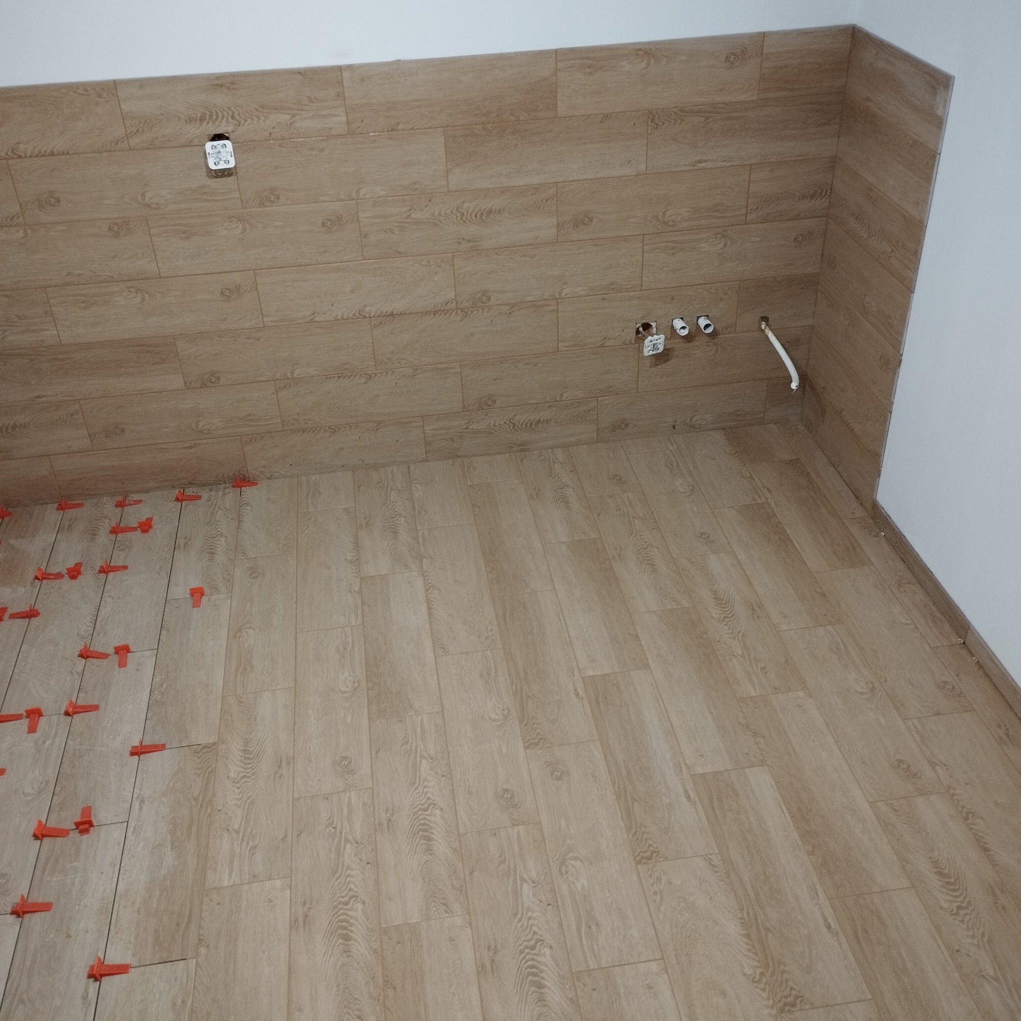 Świeżo ułożone płytki imitujące drewno na podłodze i ścianach łazienki, z widocznymi krzyżykami dystansowymi, instalacją wodną i elektryczną.