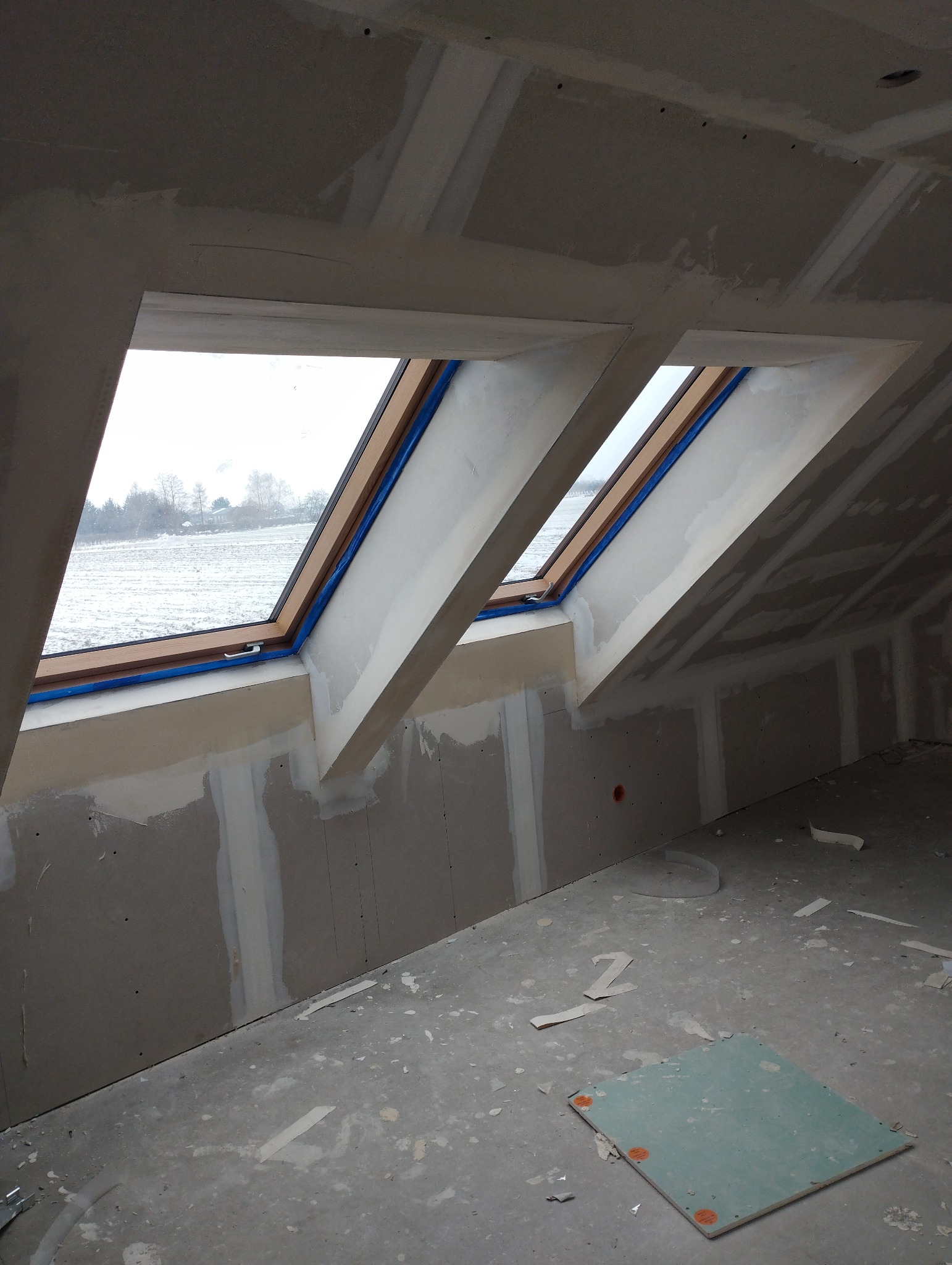 Poddasze w trakcie wykańczania: widoczne dwa okna dachowe z drewnianą ramą zabezpieczone niebieską taśmą malarską, ściany i sufit pokryte płytami kartonowo-gipsowymi, częściowo zaszpachlowane,...