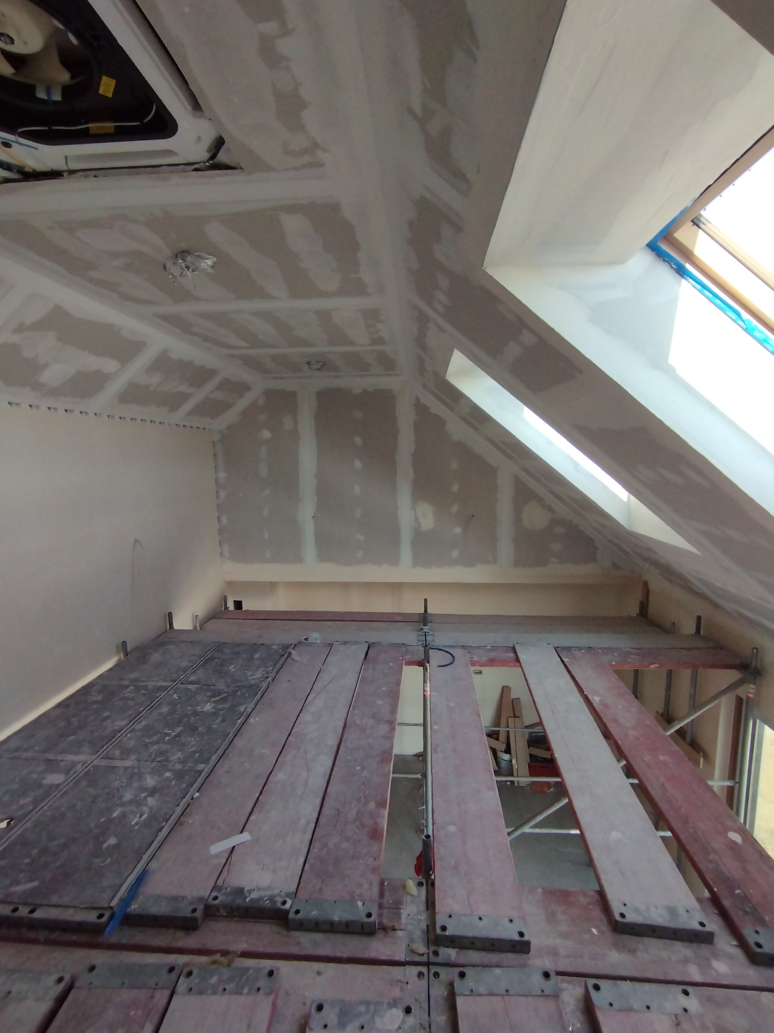 Poddasze w trakcie prac wykończeniowych, widoczne płyty kartonowo-gipsowe na skosach i suficie, częściowo zaszpachlowane, ustawione rusztowanie robocze.