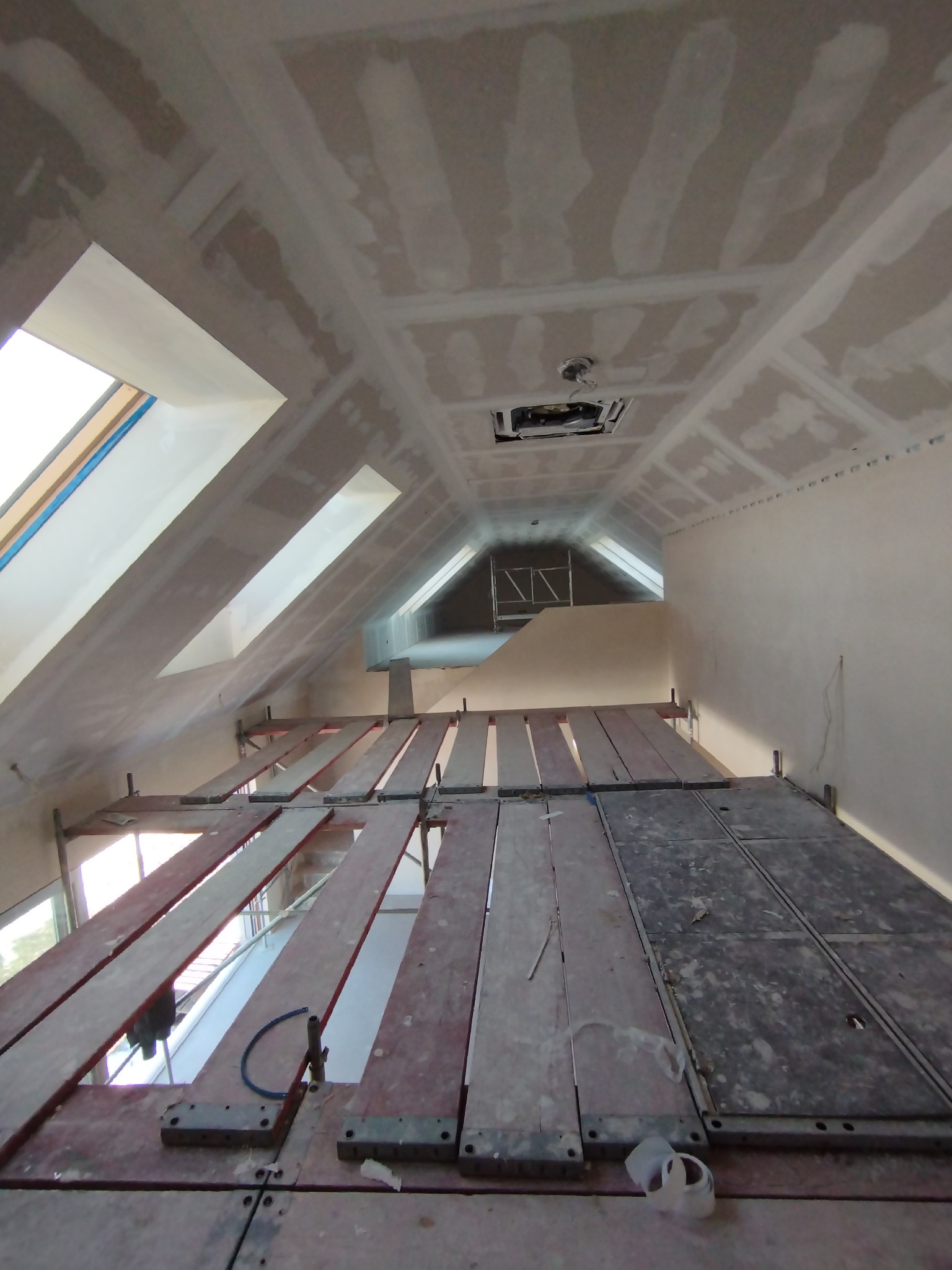Poddasze w trakcie prac wykończeniowych: szpachlowana sufit i ściany, widoczne okna dachowe, rusztowanie robocze z deskami i niebieskim przewodem.