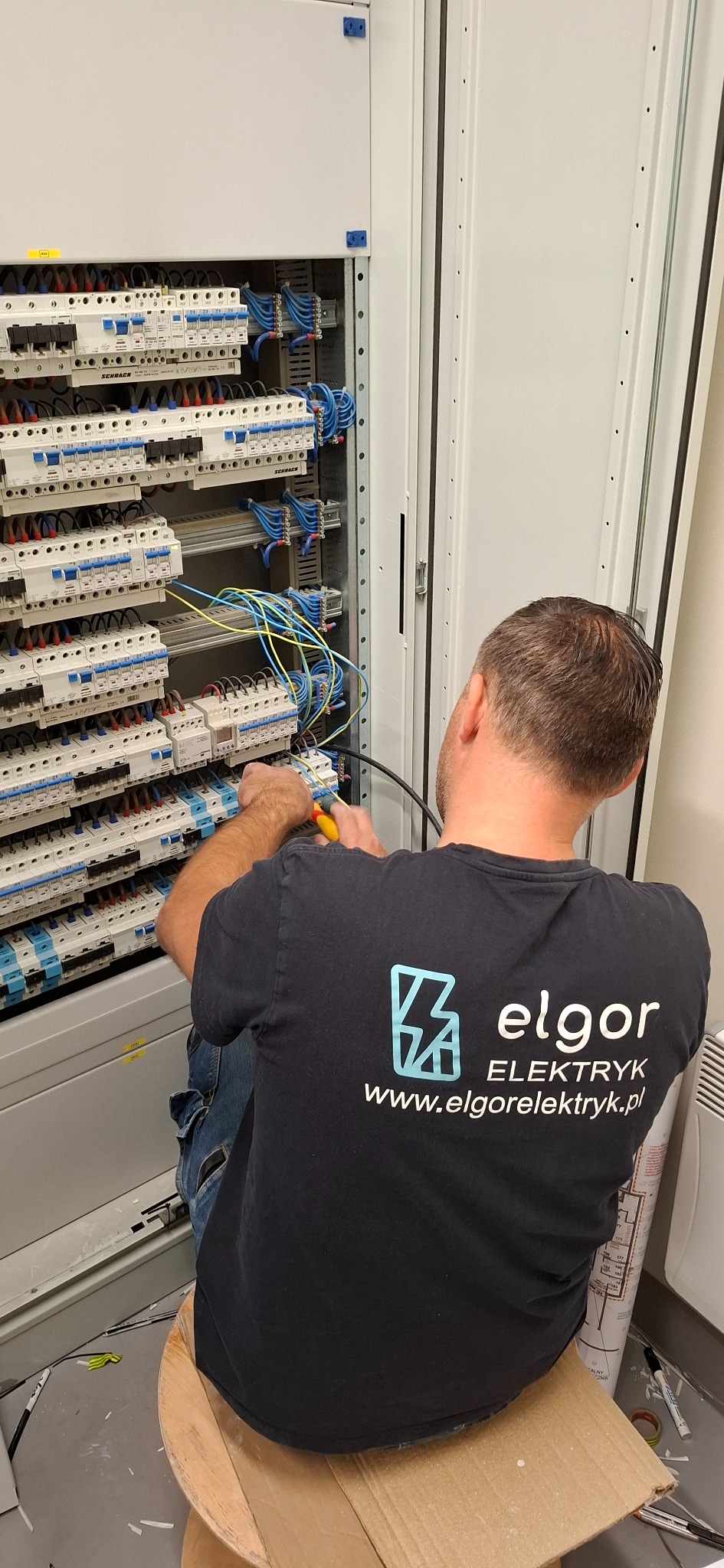 Elektryk w czarnej koszulce z logo firmy Elgor Elektryk, siedząc na kartonowym stołku, podłącza przewody w rozdzielnicy elektrycznej z licznymi bezpiecznikami i wyłącznikami, używając...