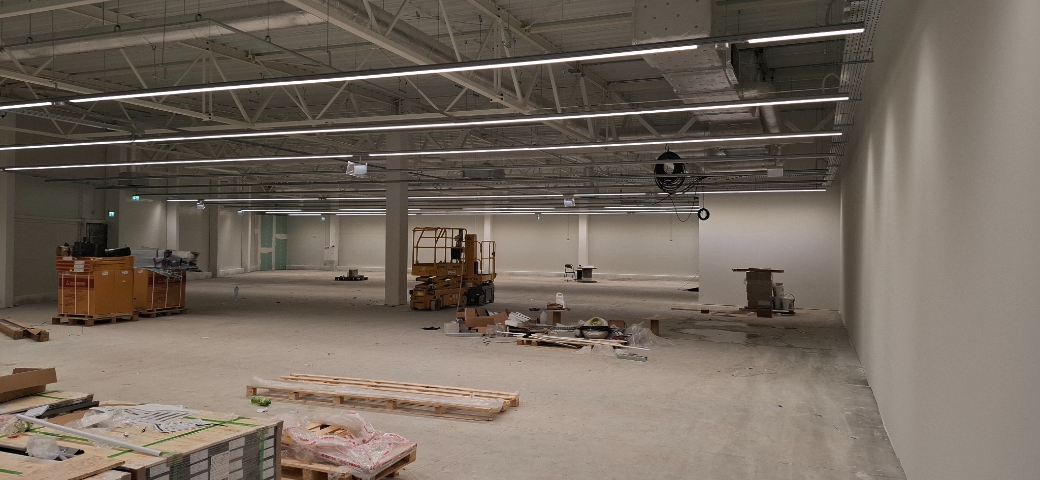 Wnętrze hali w trakcie montażu oświetlenia liniowego LED, widoczny podnośnik koszowy, palety z materiałami i narzędziami, surowy beton na podłodze.