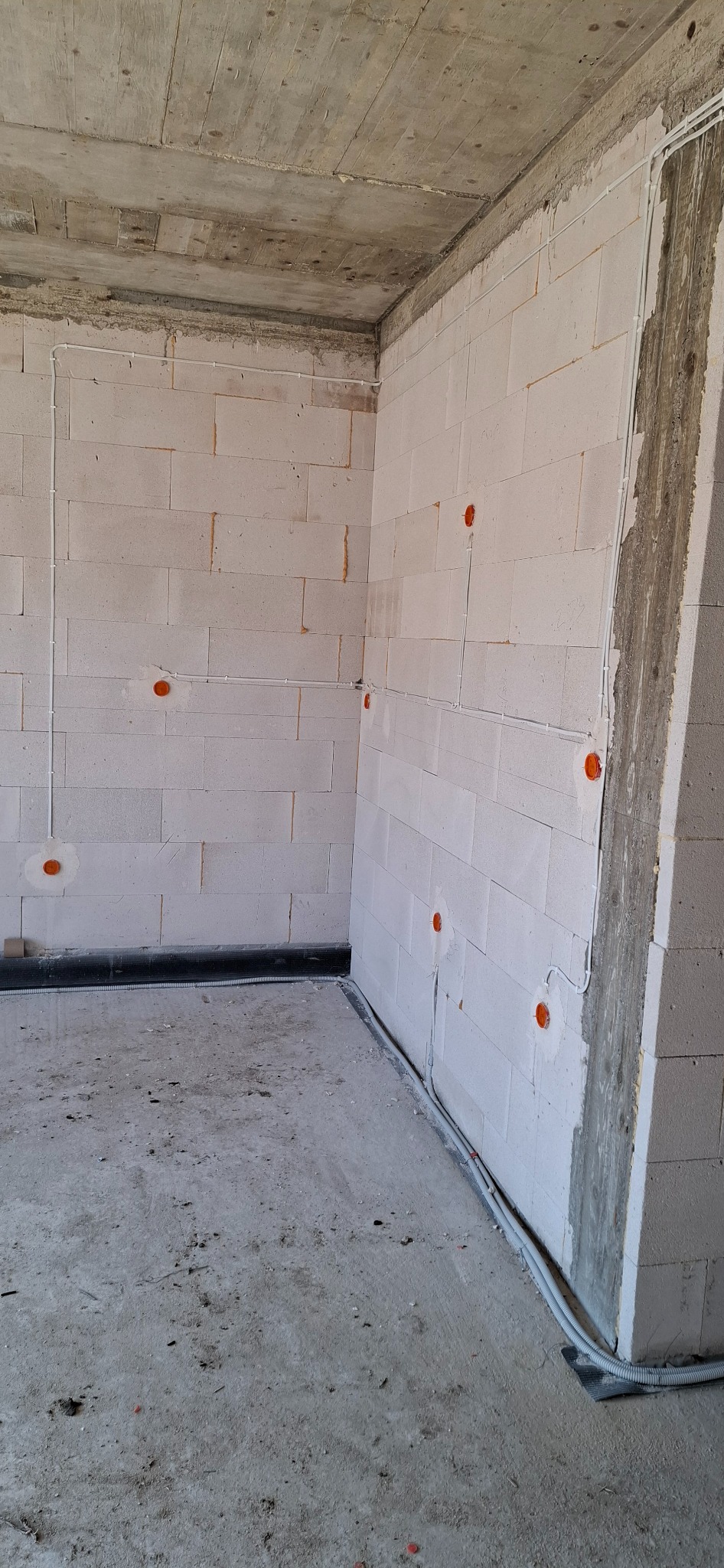 Surowe wnętrze w stanie deweloperskim z widocznymi przewodami elektrycznymi w białych rurkach na ścianach z betonu komórkowego i z pomarańczowymi puszkami instalacyjnymi, oraz rurkami przy podłodze.