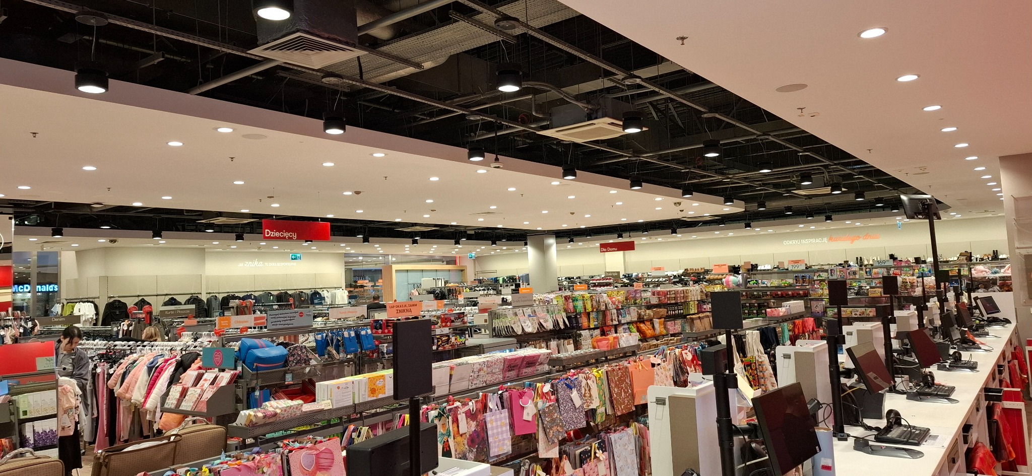 Wnętrze dużego sklepu z widoczną instalacją oświetleniową na suficie, składającą się z wielu punktowych lamp i przewodów podwieszonych pod stropem.