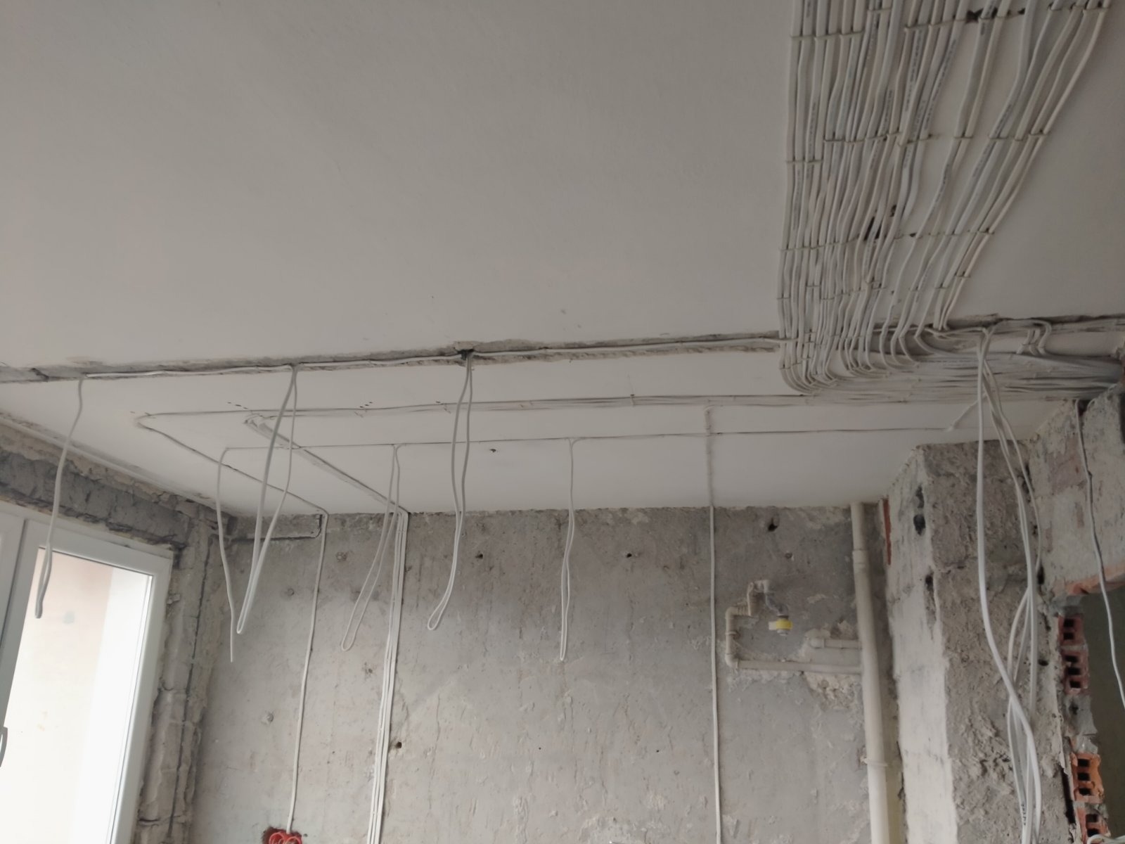 Surowy stan: białe przewody elektryczne ułożone w wiązkach pod sufitem, część przewodów zwisa luźno w dół, ściany w stanie deweloperskim z widoczną cegłą i instalacjami.