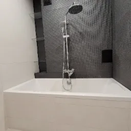 Nowoczesna łazienka z białą wanną obłożoną płytkami, ciemnoszara ściana z mozaiki z deszczownicą i półkami z przezroczystego szkła.