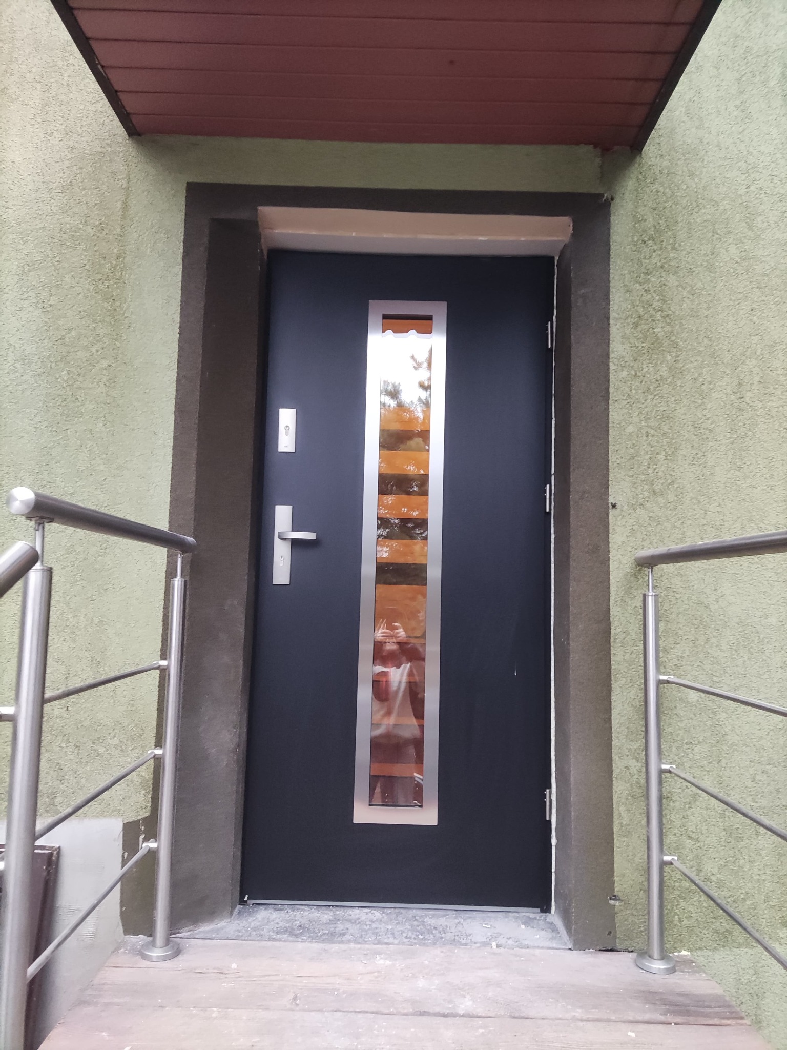 Ciemne drzwi wejściowe z podłużnym przeszkleniem, osadzone w szarozielonej elewacji, widoczne z poziomu schodów z metalową barierką.