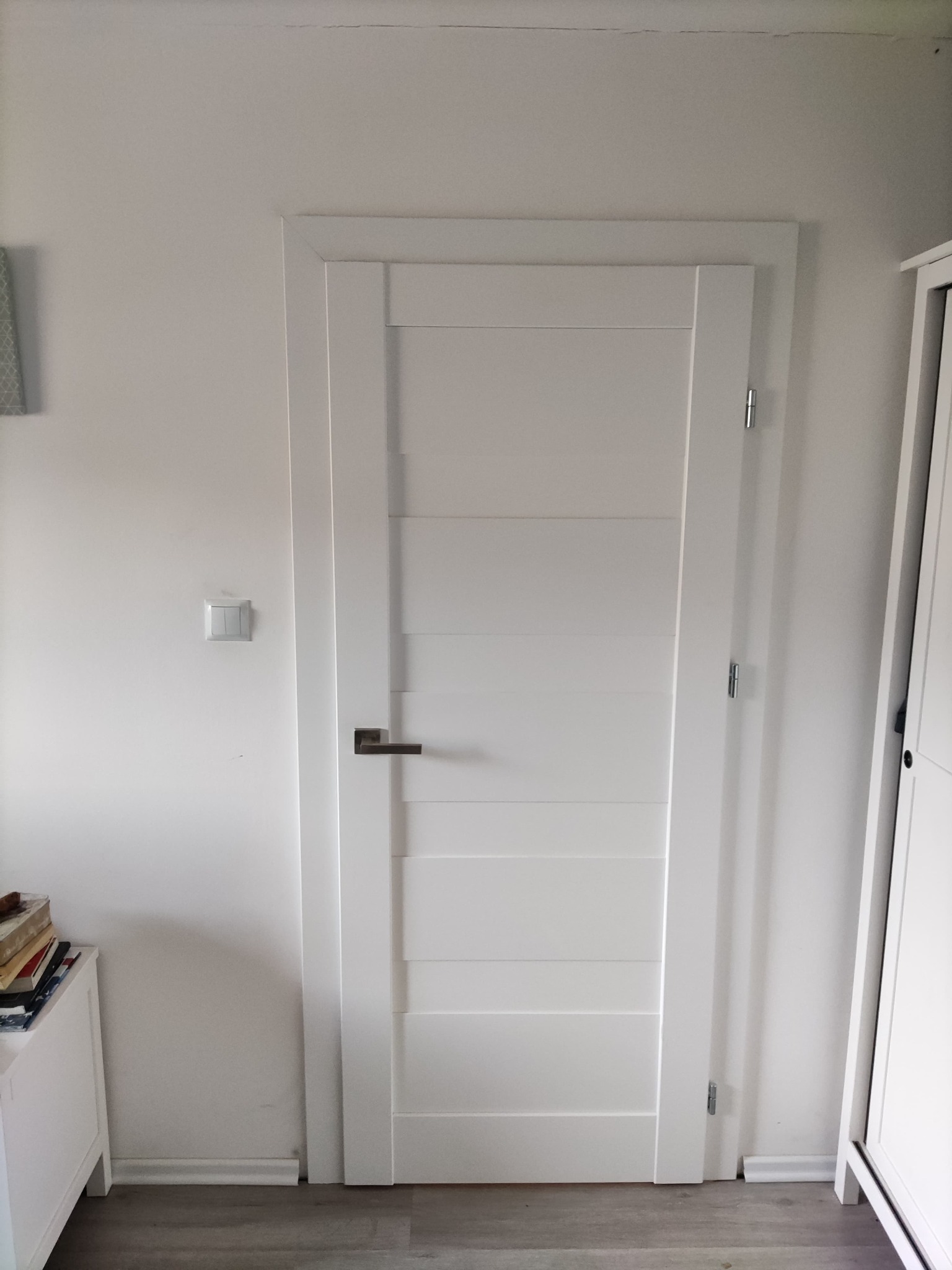 Białe drzwi wewnętrzne z poziomymi panelami, zamontowane w białej ścianie, widoczne zawiasy i klamka, fragment białej szafki po prawej stronie.