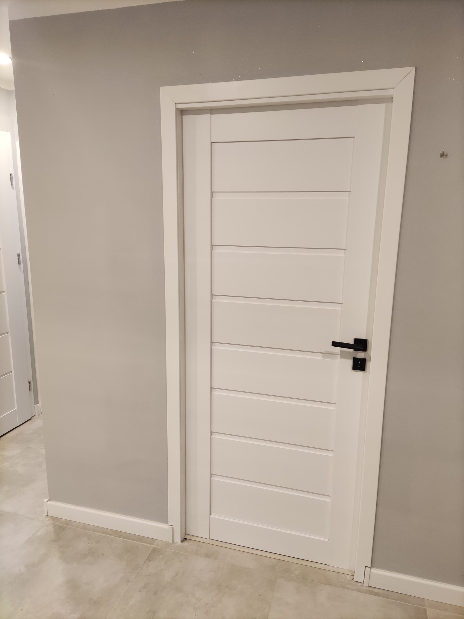 Białe drzwi wewnętrzne z poziomymi panelami i czarną klamką na tle szarej ściany i płytek podłogowych.