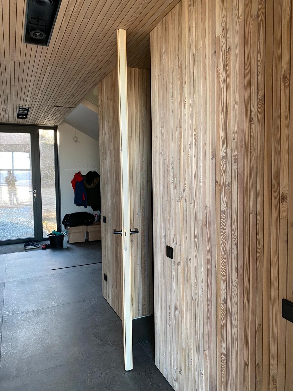 Wnętrze z drewnianym wykończeniem ścian i sufitu, widoczne otwarte drzwi wewnętrzne z czarną klamką i fragment wejścia z widoczną osobą w odbiciu szyby.