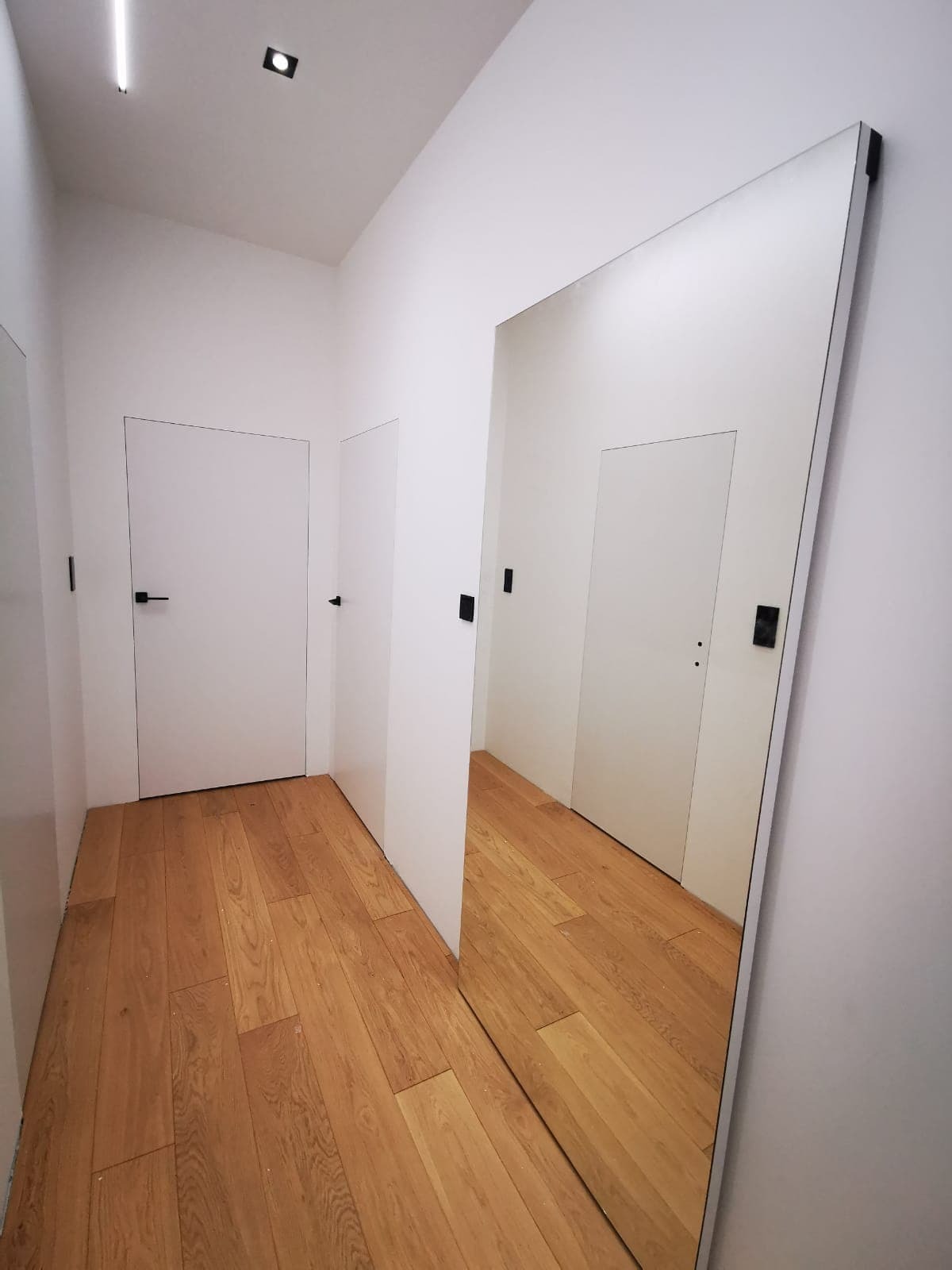 Wnętrze korytarza z drewnianą podłogą, białymi ścianami i ukrytymi drzwiami z czarnymi klamkami, duże lustro na ścianie odbija widok korytarza.