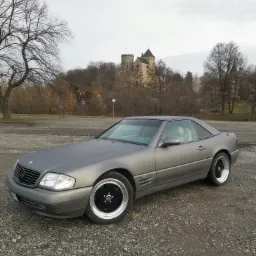 Matowy szary Mercedes SL z czarnymi felgami na żwirowym parkingu, zamek w tle.