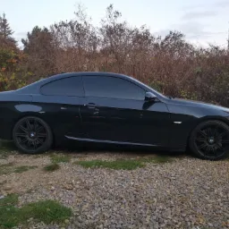 Ciemny samochód BMW z przyciemnionymi szybami, zaparkowany na żwirowym podłożu w Będzinie, na tle krzewów i drzew.