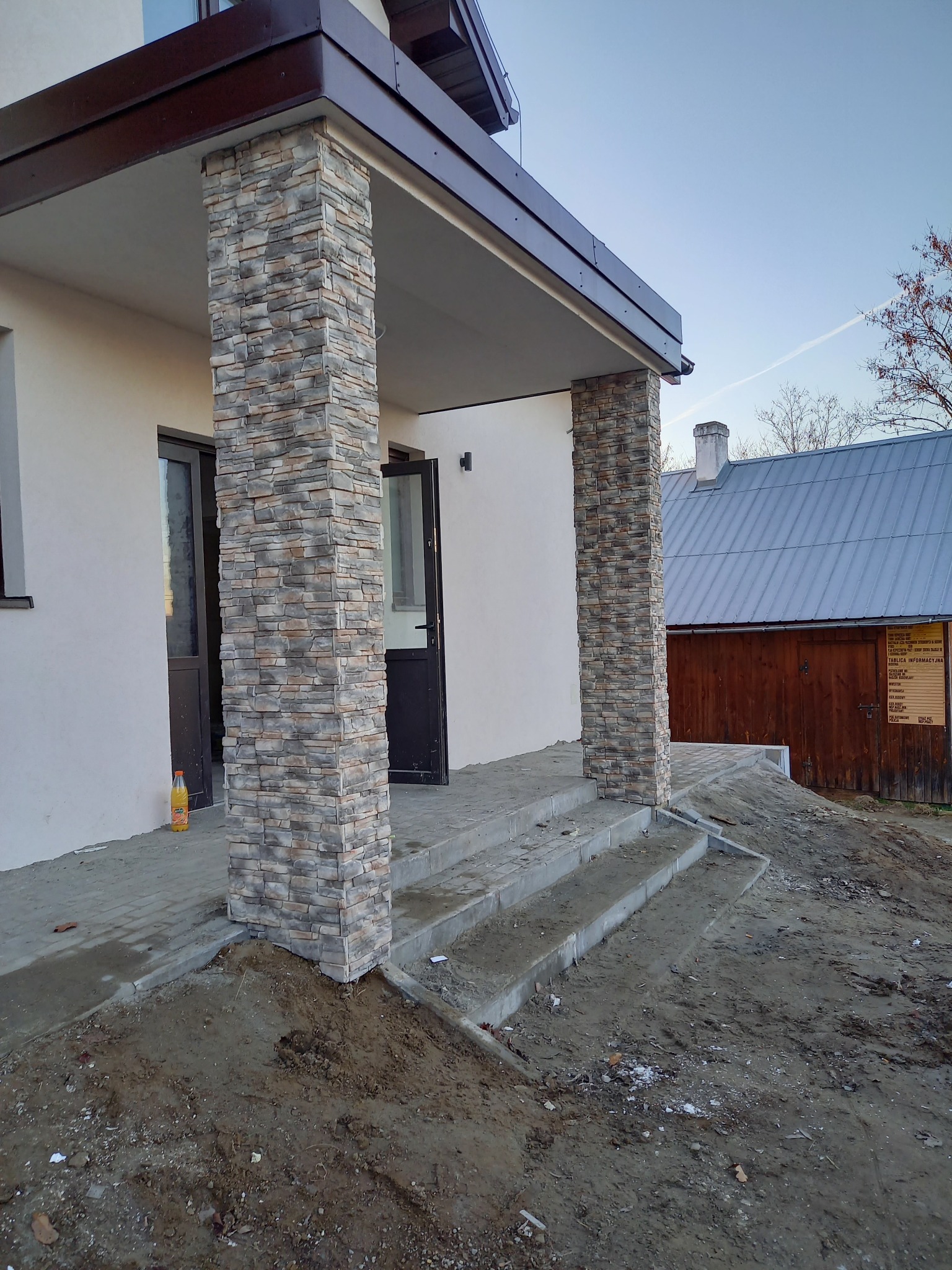 Wejście do domu z kolumnami wykończonymi kamieniem dekoracyjnym, widoczne schody wejściowe i częściowo zagospodarowany teren wokół budynku.