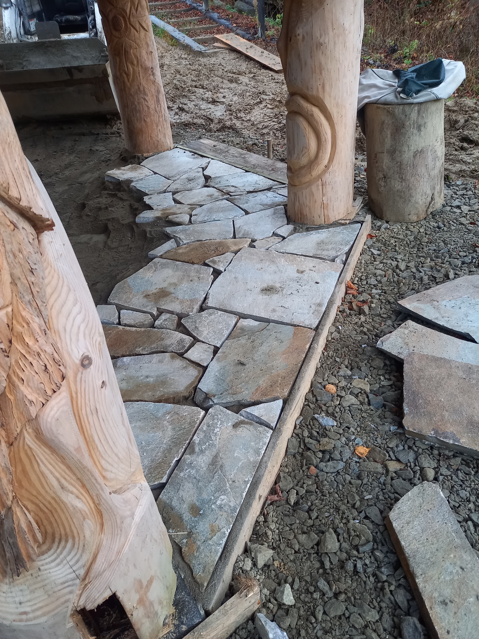 Nieregularne kamienne płyty tworzące ścieżkę, otoczone drewnianymi słupami z ręcznie rzeźbionymi wzorami, w trakcie układania na żwirowym podłożu.