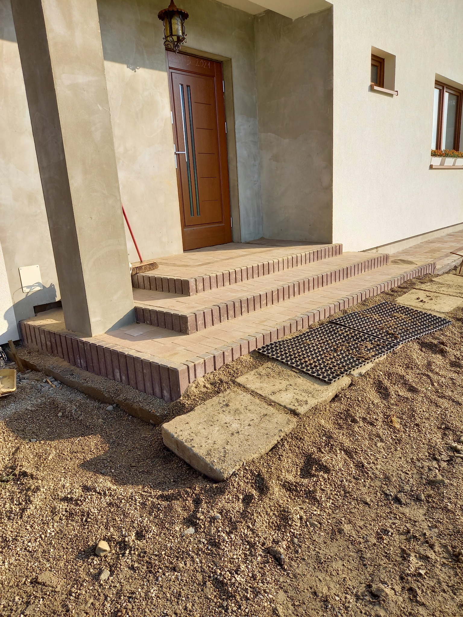 Nowe schody wejściowe z cegły klinkierowej prowadzące do drzwi wejściowych w domu jednorodzinnym; widoczny fragment elewacji i lampa nad drzwiami.