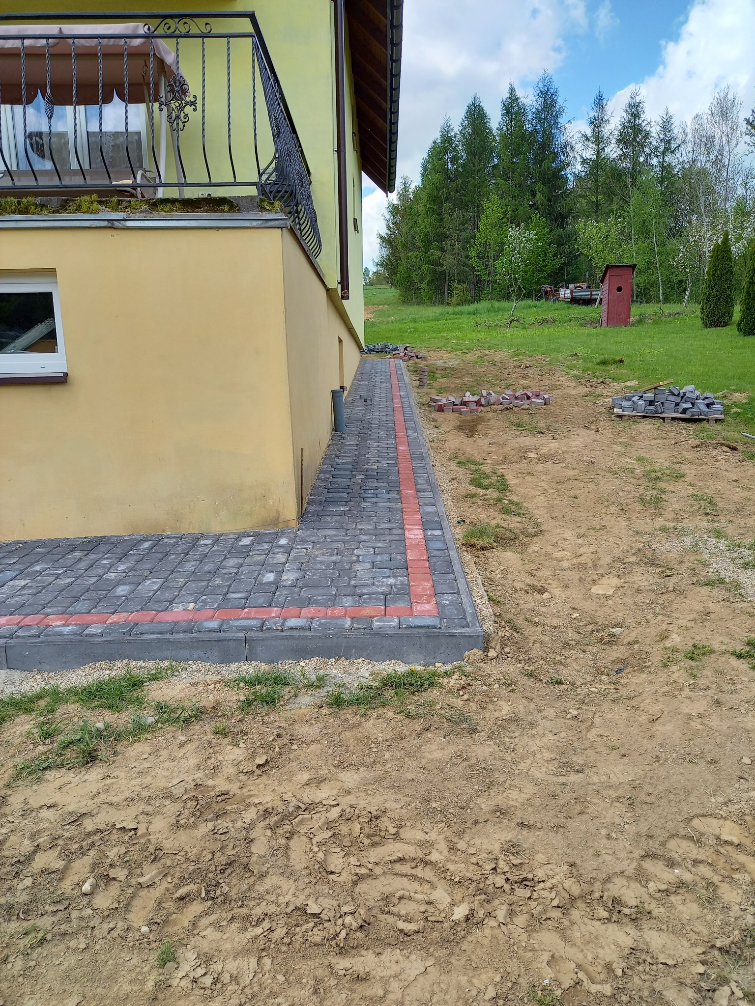 Ścieżka z kostki brukowej w kolorze szarym z czerwonym obramowaniem, prowadząca wzdłuż żółtej ściany budynku, z widocznymi stertami kostki brukowej na ziemi i zielenią w tle.