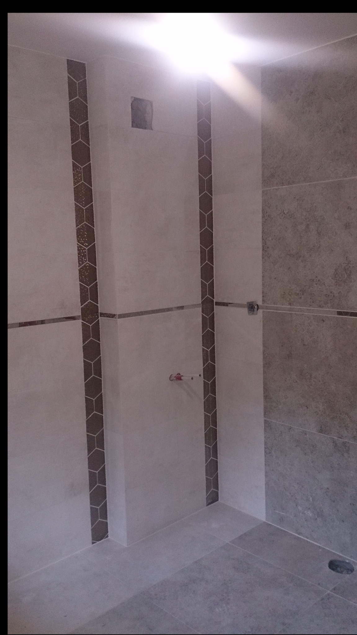 Fragment łazienki w trakcie wykańczania, widoczne pionowe pasy dekoracyjne z motywem heksagonów na jasnych płytkach, surowy otwór na instalację elektryczną oraz szare płytki imitujące kamień po...