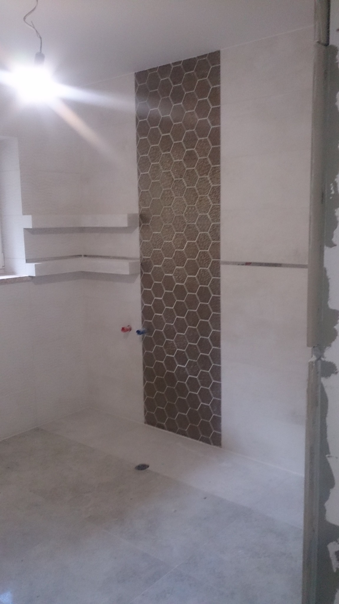 Wnętrze łazienki w trakcie prac wykończeniowych, widoczna mozaika w kształcie plastra miodu na ścianie, świeżo położone płytki podłogowe, wystające rurki instalacji wodnej i półki w narożniku